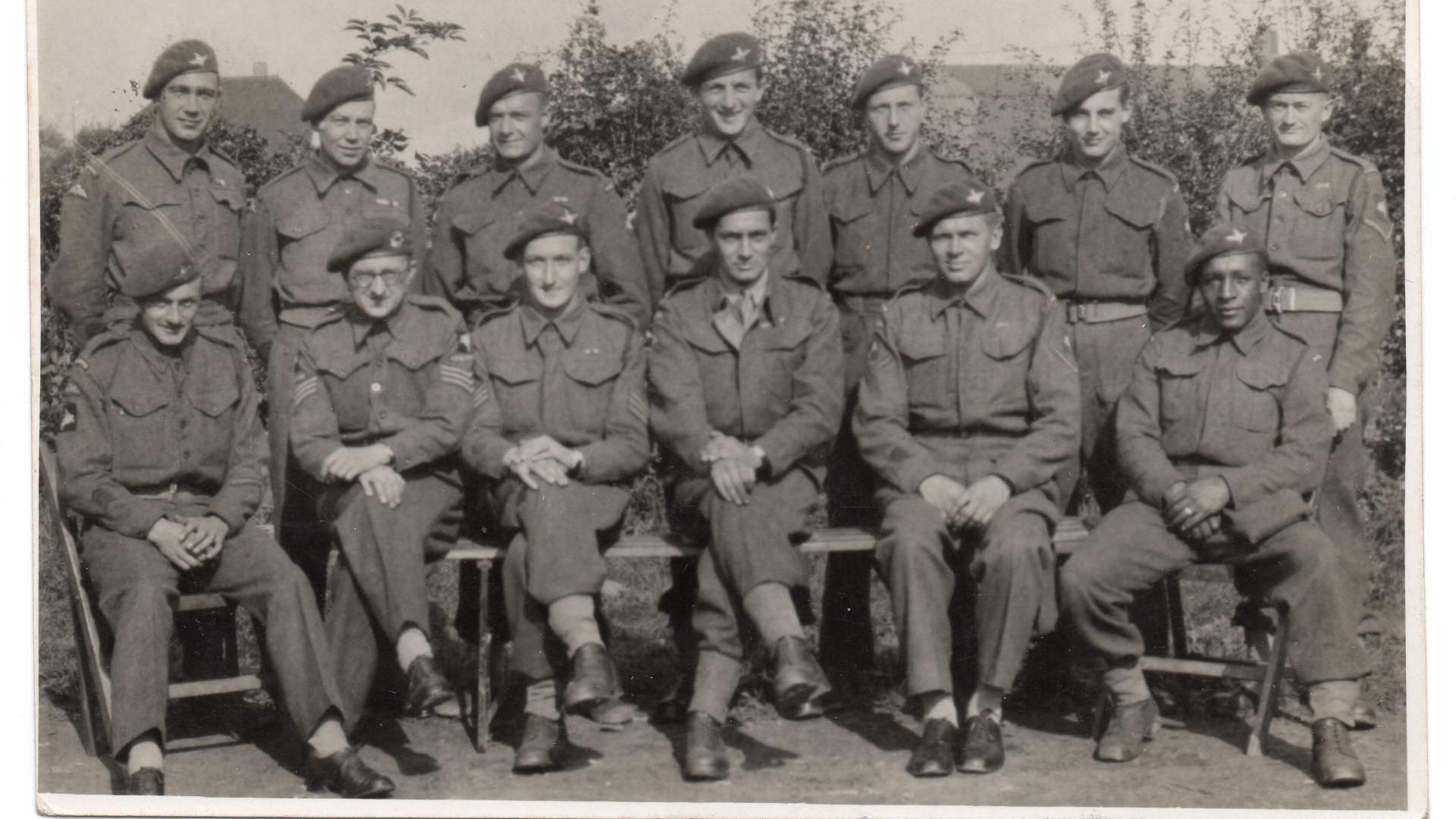 A black and white photo of soldiers wearing suits and berets. Six men are sitting on the front row  in one row and seven are standing at the back.