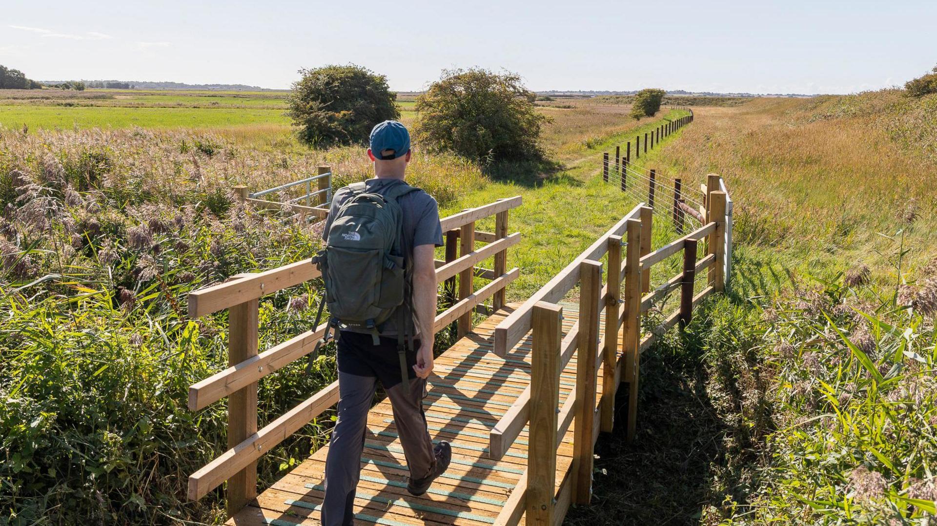 A man walks across a small wooden bridge, he is wearing a blue tshirt, trousers, a baseball cap and a backpack. Around the bridge is foliage, with fields in the background. 