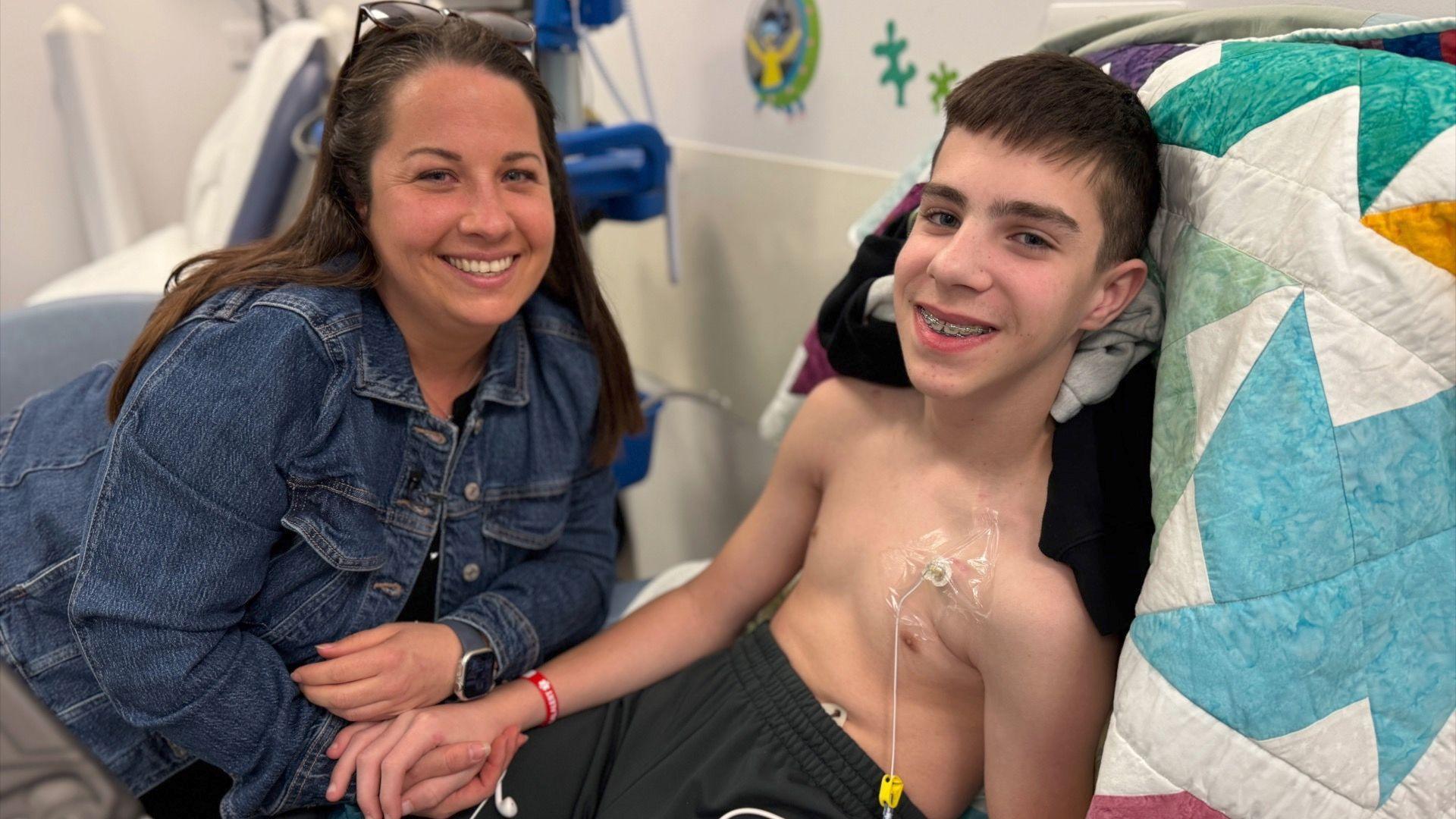 Finley and his mum Rhys in a hospital room in Broomfield Hospital. Finley is lying upright on the hospital bed, he is not wearing a top as he has a medical tube running into his chest. He is lying on top of a multi-coloured patchwork blanket, and he wears black tracksuit bottoms. He is holding hands with his mum Rhys who is sat beside him. Rhys has a denim blue jacket on and has dark brown hair running down past her shoulders. She has a pair of sunglasses sitting on top of her head, and both are smiling at the camera