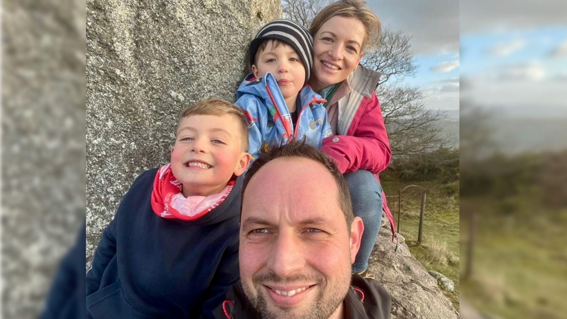 Family photo taken as a selfie. It is an outdoor photo with a rock in the background. Rhys Jenkins is in the centre and front, Ioan is on the left, behind him is young son Gruff and at the back is Rhys' wife Carrie Jenkins.