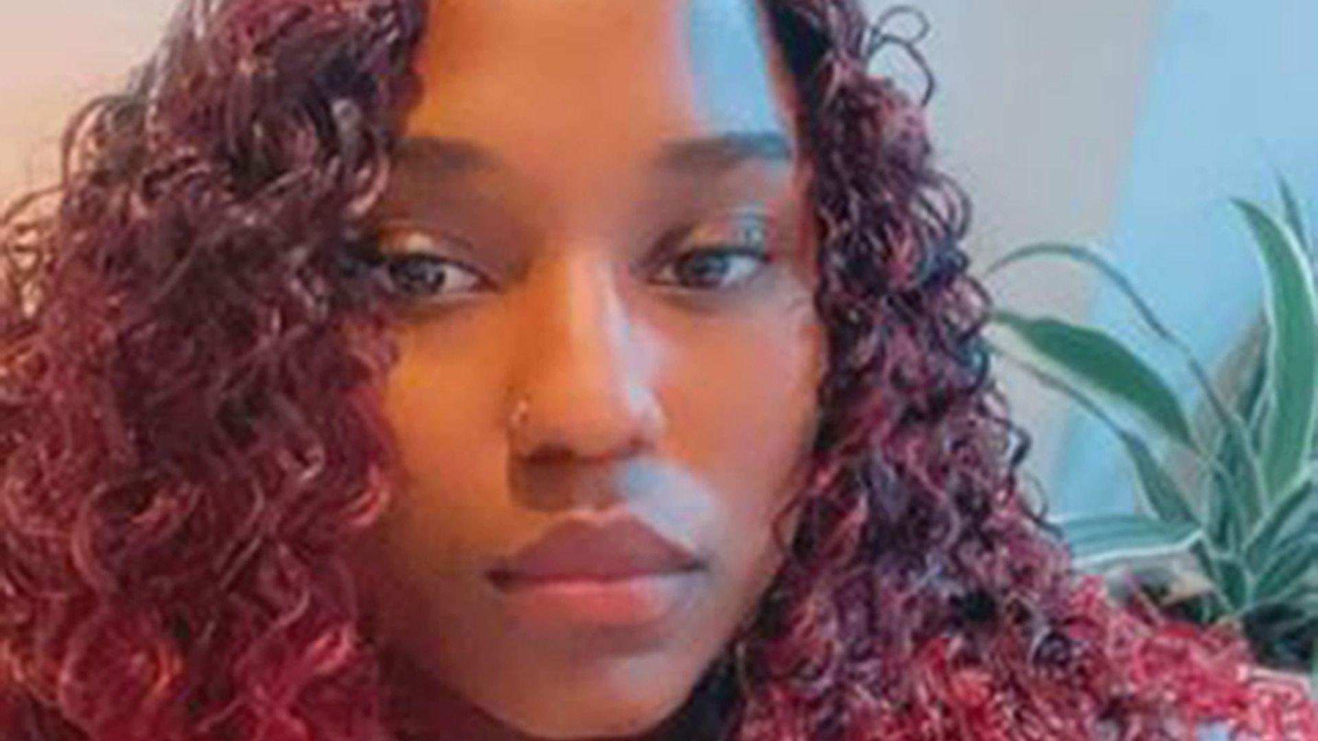 A woman with long red curly hair next to a green plant. She is looking into the camera