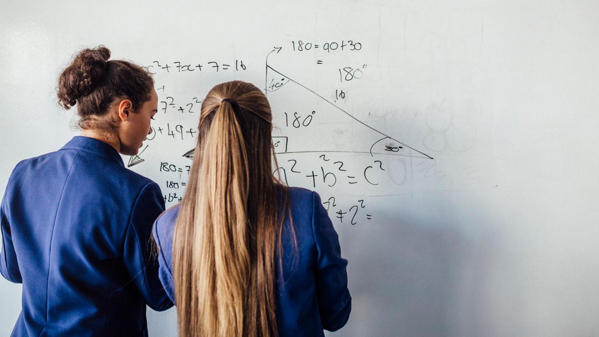 Two pupils wearing navy blue blazers face away from the camera as they write algebra on a whiteboard. The student on the left has her brown curly hair in a bun, and her classmate on the right has long light brown hair, half tied up.