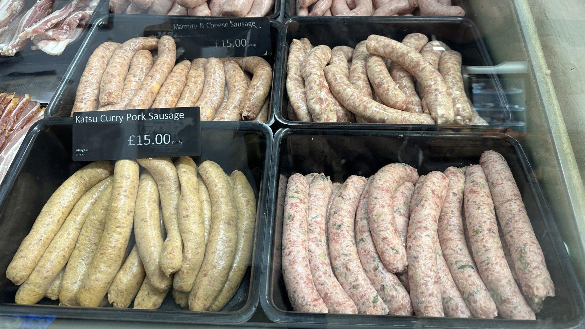 Four black plastic trays of sausages in a butcher's display. The sausages vary slightly in colour by variety. Two of them are labelled "katsu curry pork sausage" and "Marmite and cheese sausage", both at £15/kg.