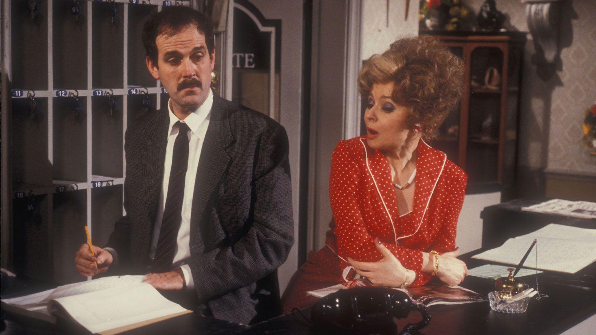 Prunella Scales in a red dress leaning on the reception desk as Sybil in Fawlty Towers and John Cleese as Basil