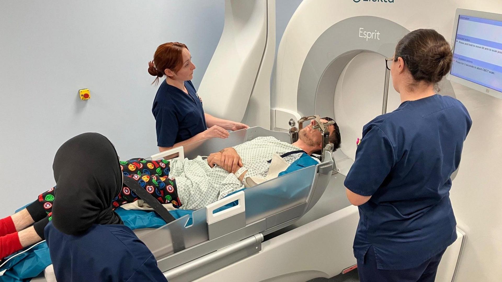 A man lying down on a machine about to go in to have radiation treatment on his brain. He has three female nurses in blue uniforms around him.