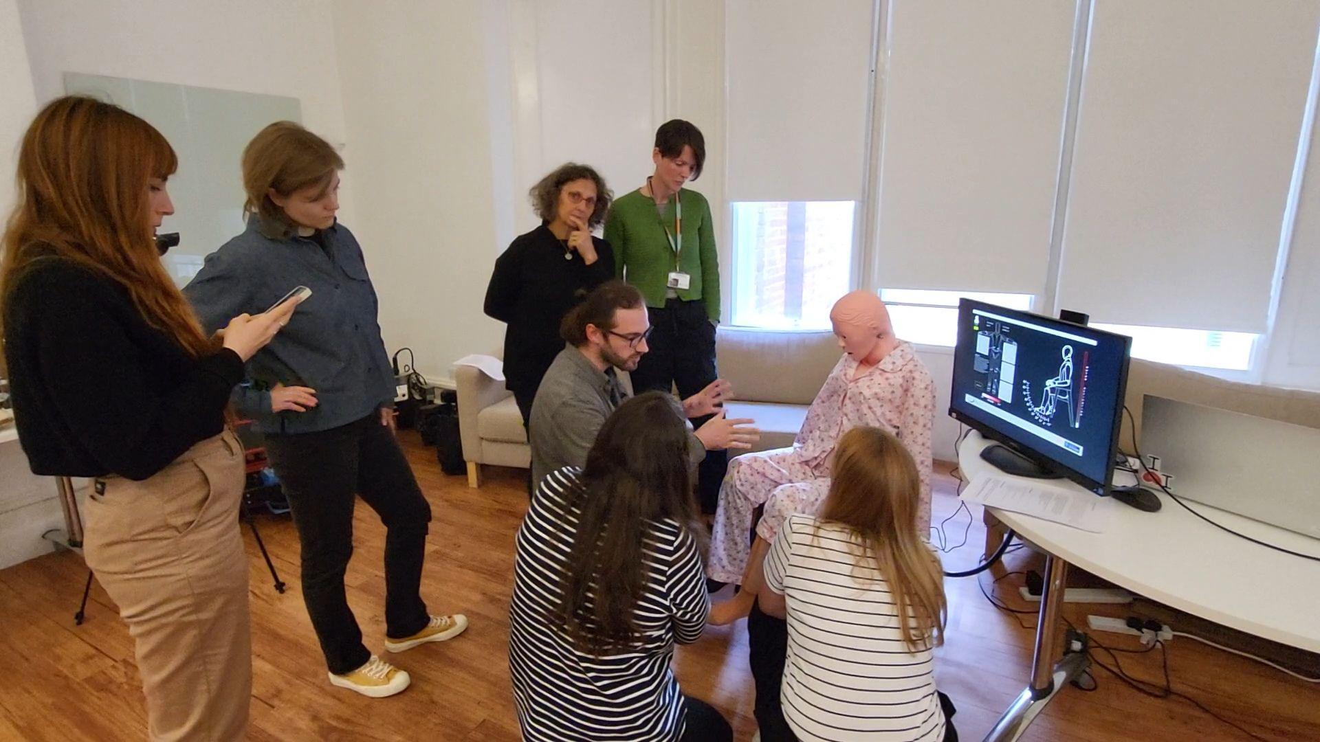 The research team gathered around Mona the robot who is dressed in a pink pyjamas. Some of the researchers are taking notes, others are listening to a colleague who is talking to them while kneeling in front of the robot. Two other researchers are kneeling next to him. There is a monitor showing the results of Mona's mobility.