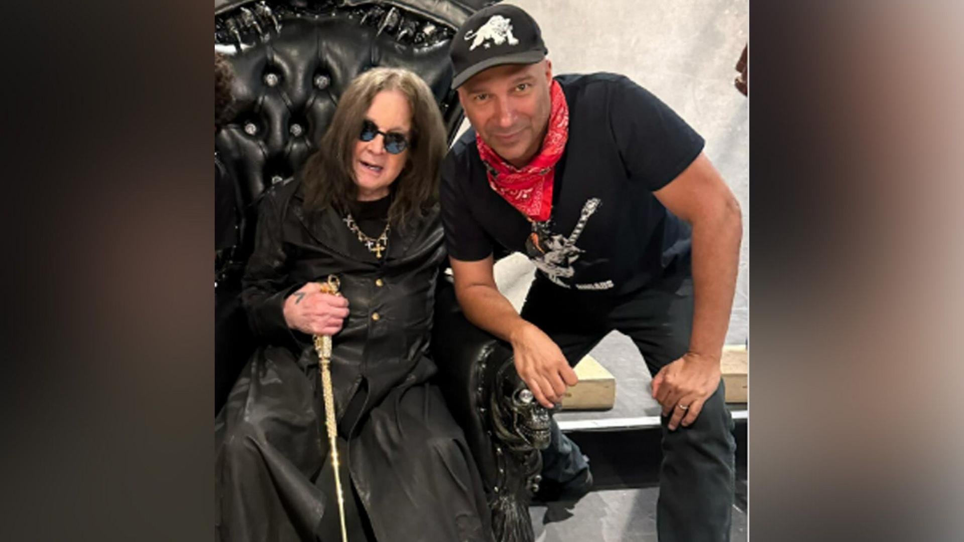 Ozzy Osbourne in a black trenchcoat sits in a black leather throne while holding a gold cane. Tom Morello is crouched next to him, wearing a black t-shirt, black baseball cap and black jeans and a red neckerchief