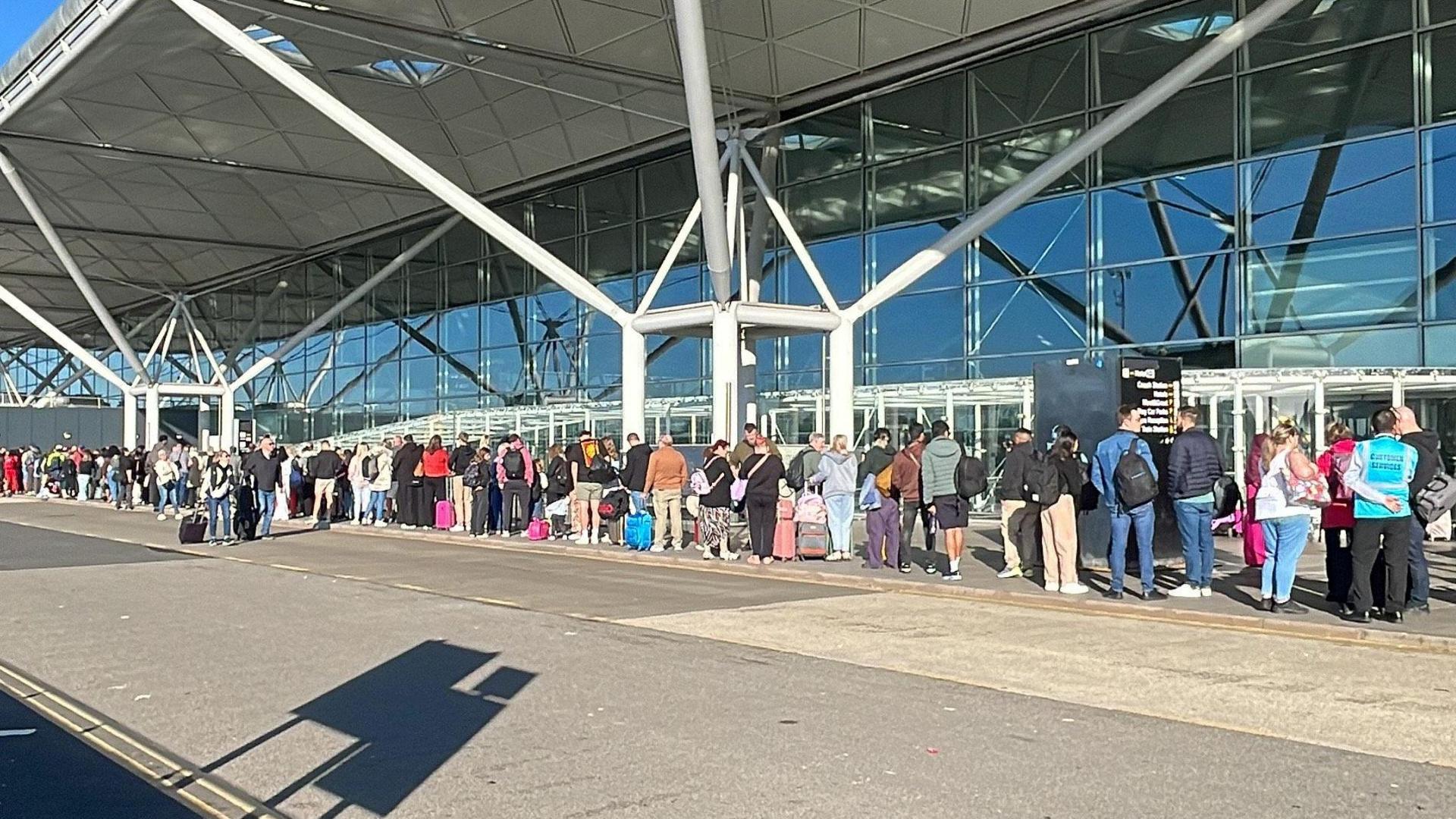 A queue of people outside an airport terminal. The sun is shining and people are carrying luggage.