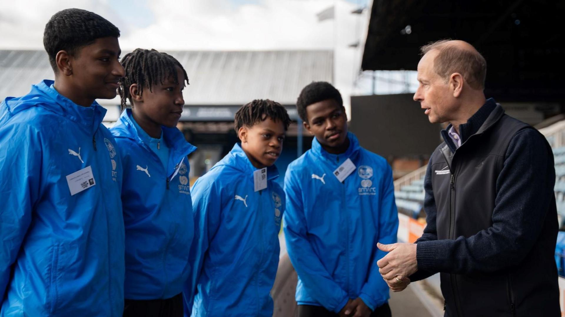 Four youngsters wearing mid-blue casual sports jackets talk to Prince Edward who is wearing a black sport jacket. They are in a football stadium.