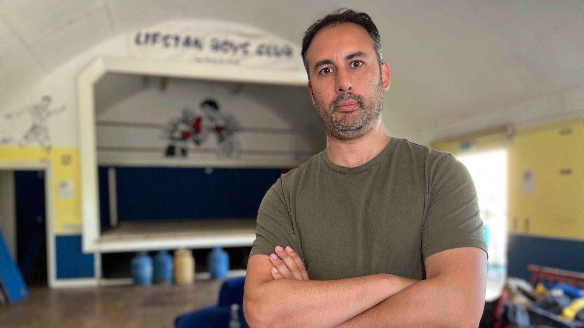 Jay Solder stands inside the Lifstan Boys Club building. He wears a dark green T-shirt, has dark short hair and a stubbly beard. He stands with his arms folded across his chest. Behind him is a stage with a boxing ring in the middle, and above the stage are the words "Lifstan Boys Club"