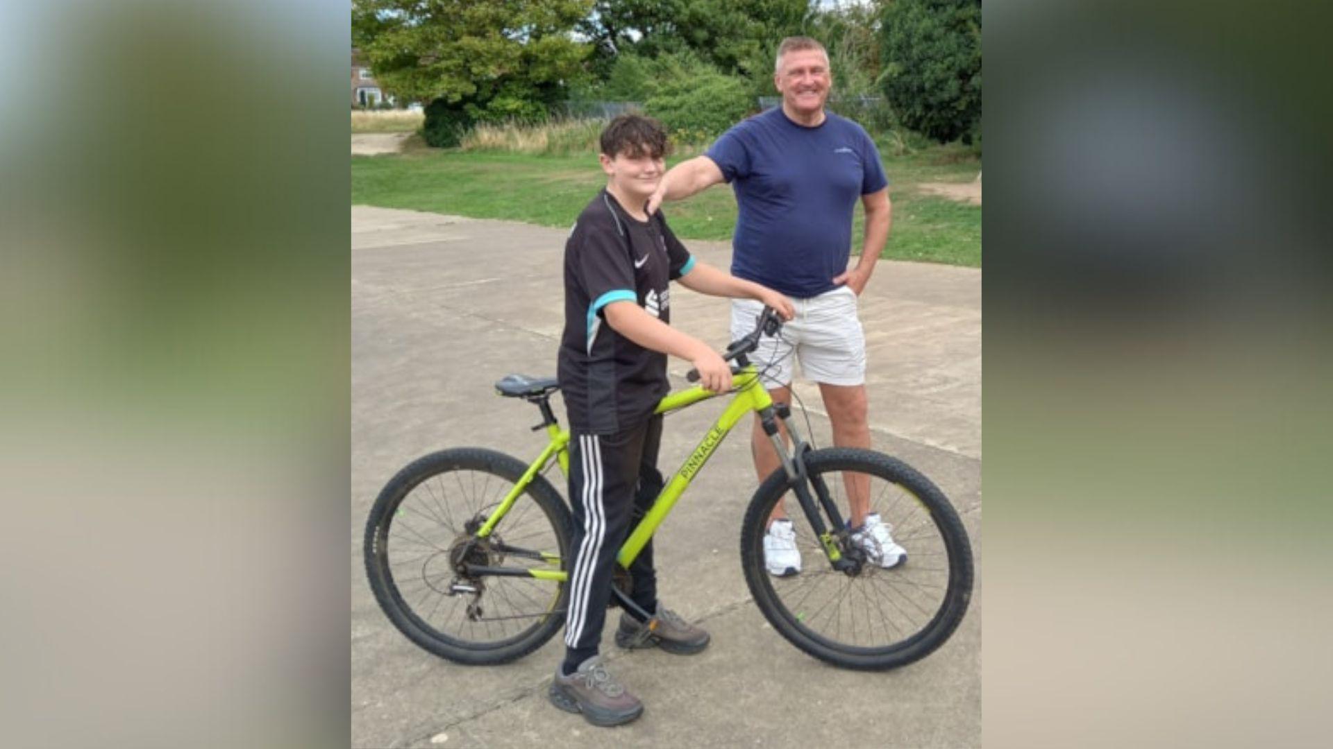 A boy wearing a black football shirt and black tracksuit bottoms with three white stripes down the side on a light green bike in a park. He is standing next to a man wearing a blue T-shirt, cream shorts and white trainers.