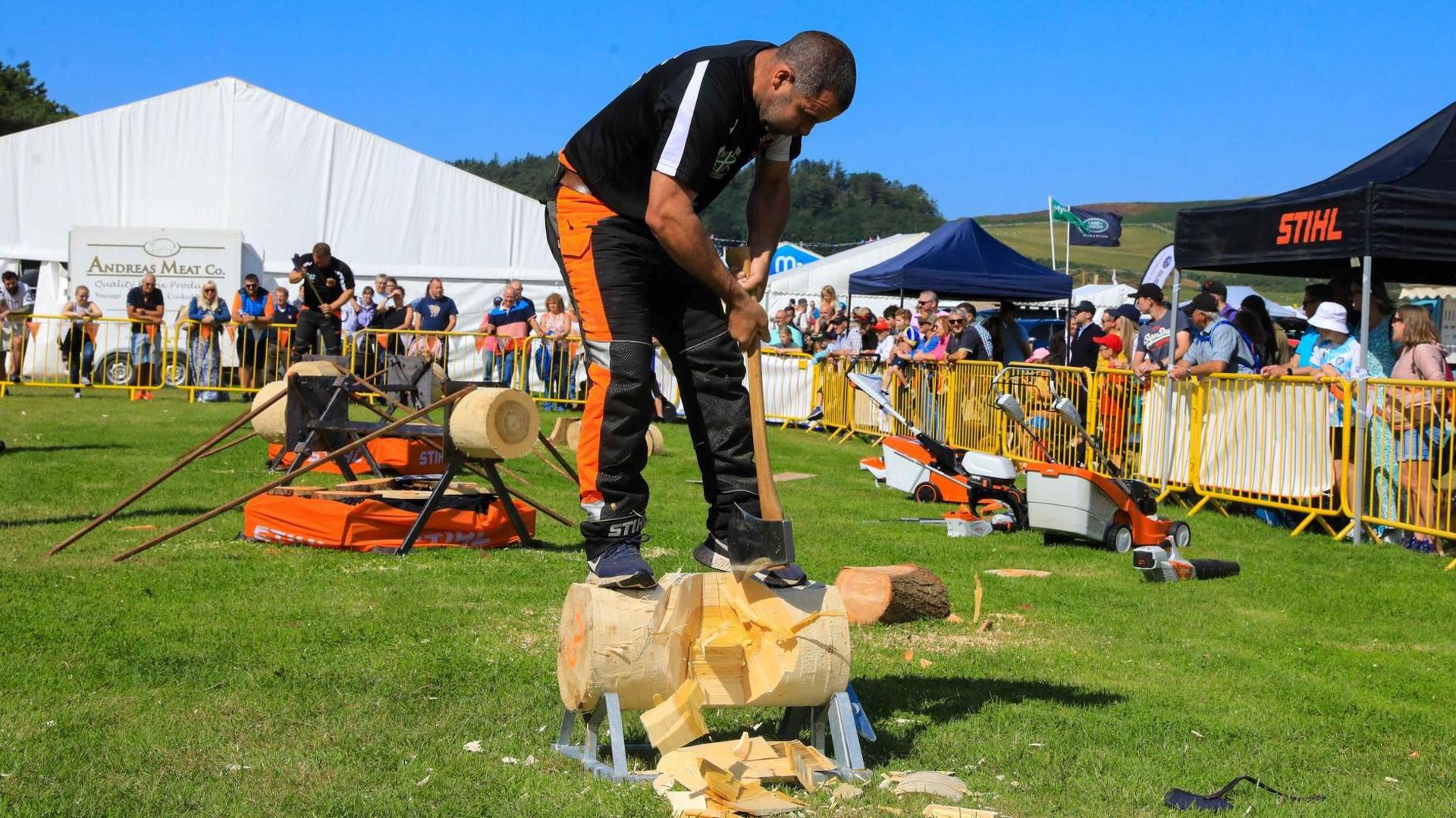 A man standing on part of a log carving it with an axe in the show ring, which has yellow barriers around it.