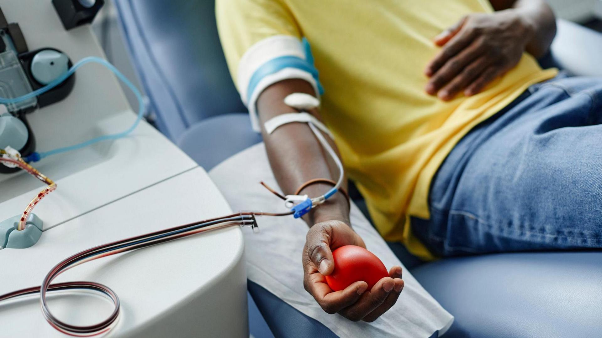 A man wearing a yellow shirt lies on a couch giving blood with a tube attached to his arm, while holding a soft red ball in his hand 