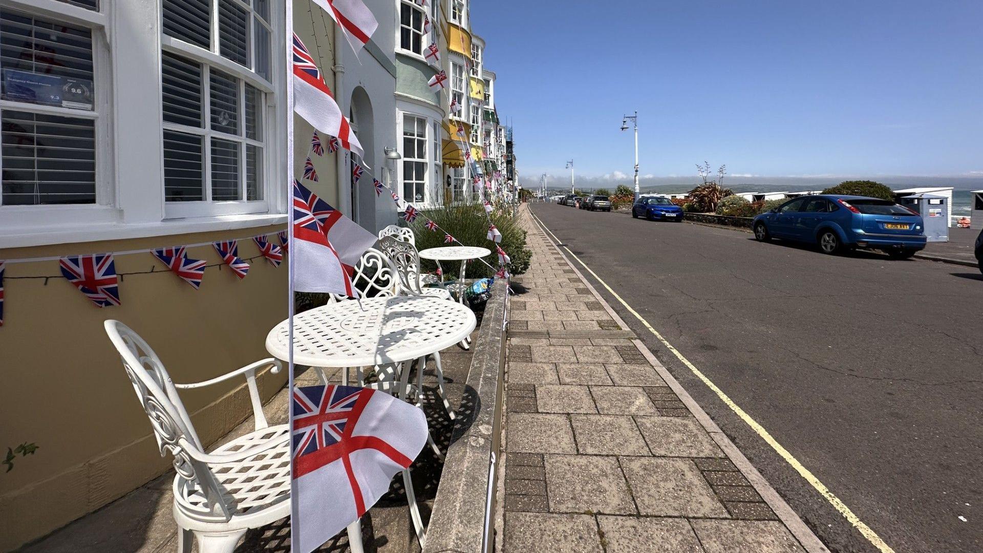 View from the pavement along a row of colourful Georgian townhouses on Weymouth seafront. The nearest building is painted yellow and has white tables and chairs outside and is decorated with bunting of the Royal Navy White Ensign flag. Cars are parked on the other side of the road and behind the cars is the beach and the sea.