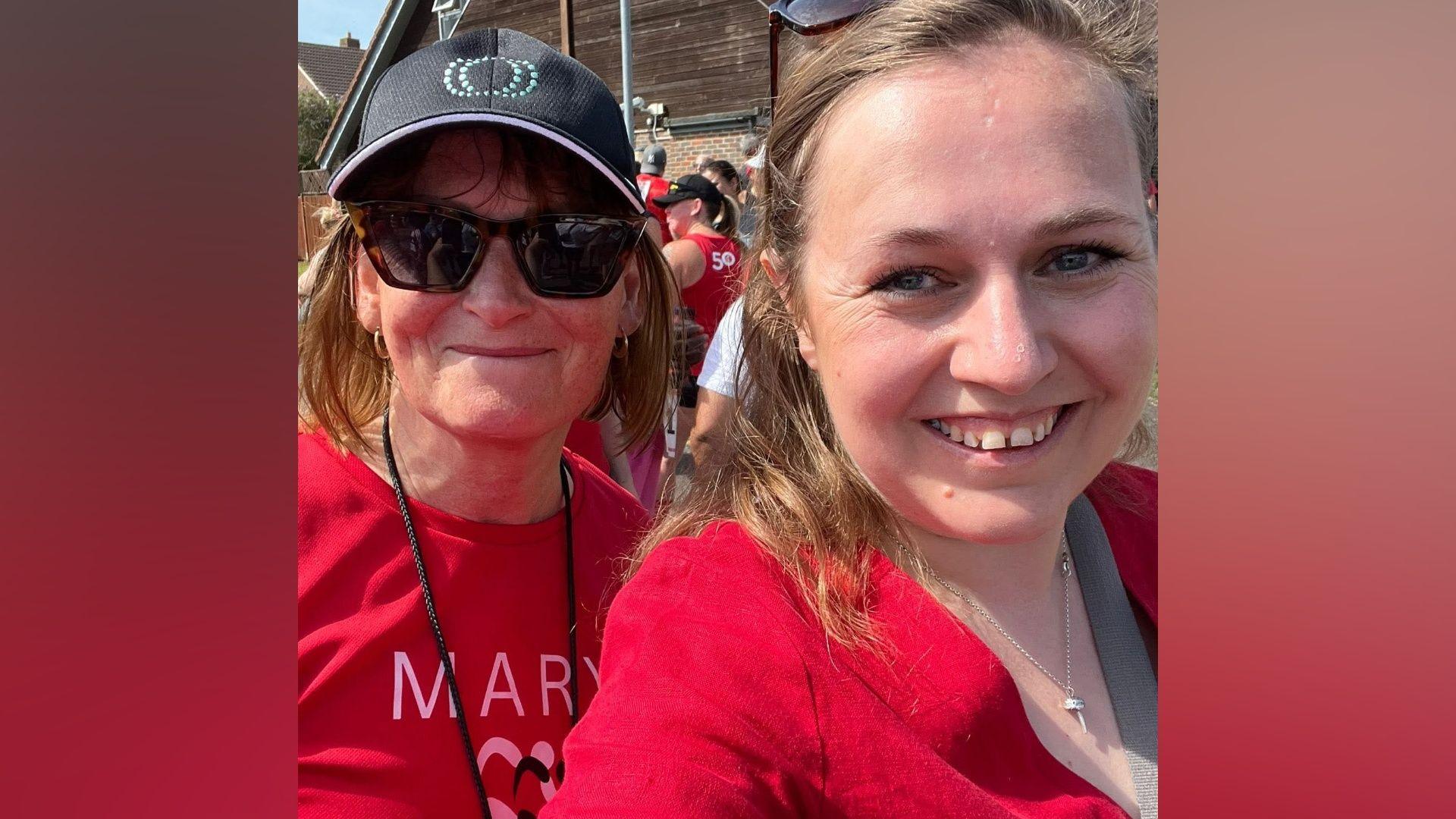Two women both wearing red; the woman on the left is wearing sunglasses and a sun hat. She has short brown hair and smiling at the camera. Another woman has blonde hair and is also wearing a red top and a small silver necklace.
They are both smiling at the camera 
