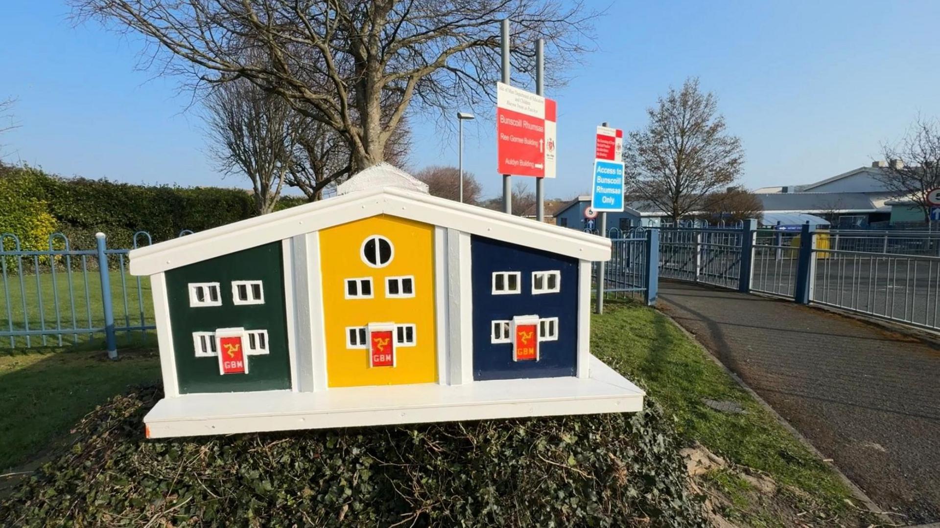 The fairy house which its white, with yellow, green and blue panels sits on a tree stump covered in green plants. It is next to the gates of a school, which is a large building in the distance. There are two red and white signs nearby with the school's name.