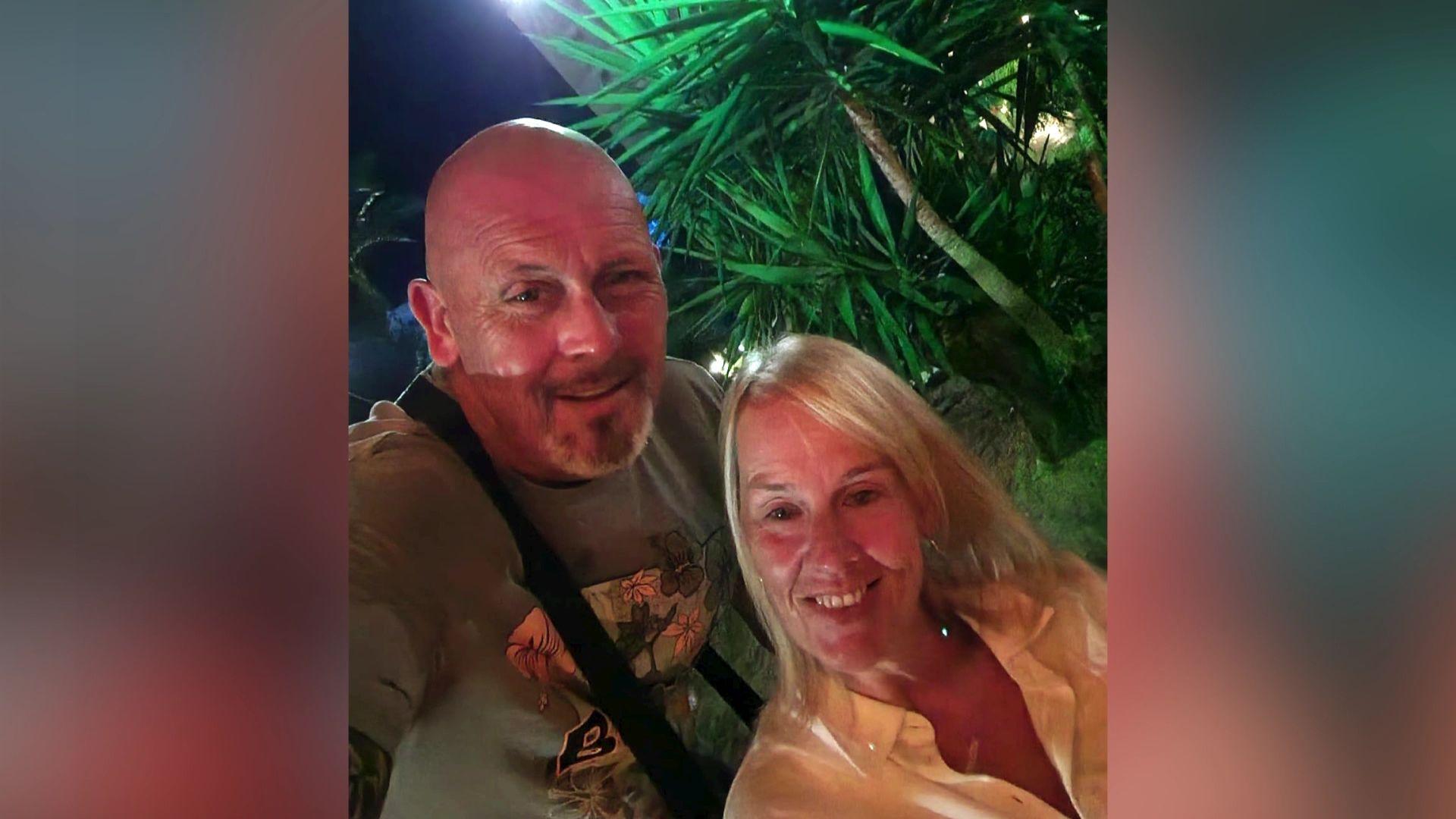 A man and a woman smile at the camera. There is a tree behind them. The man is bald and the woman has blonde hair.