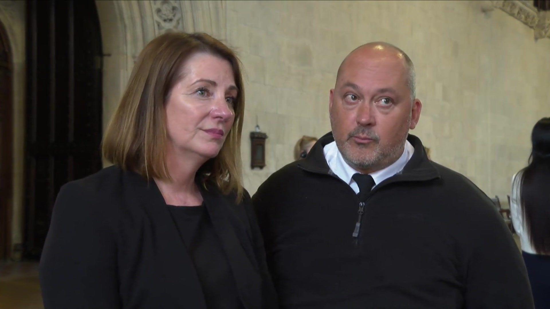 A brunette woman and a bald man from the torso up both wearing black, stood in front of a neutral coloured wall