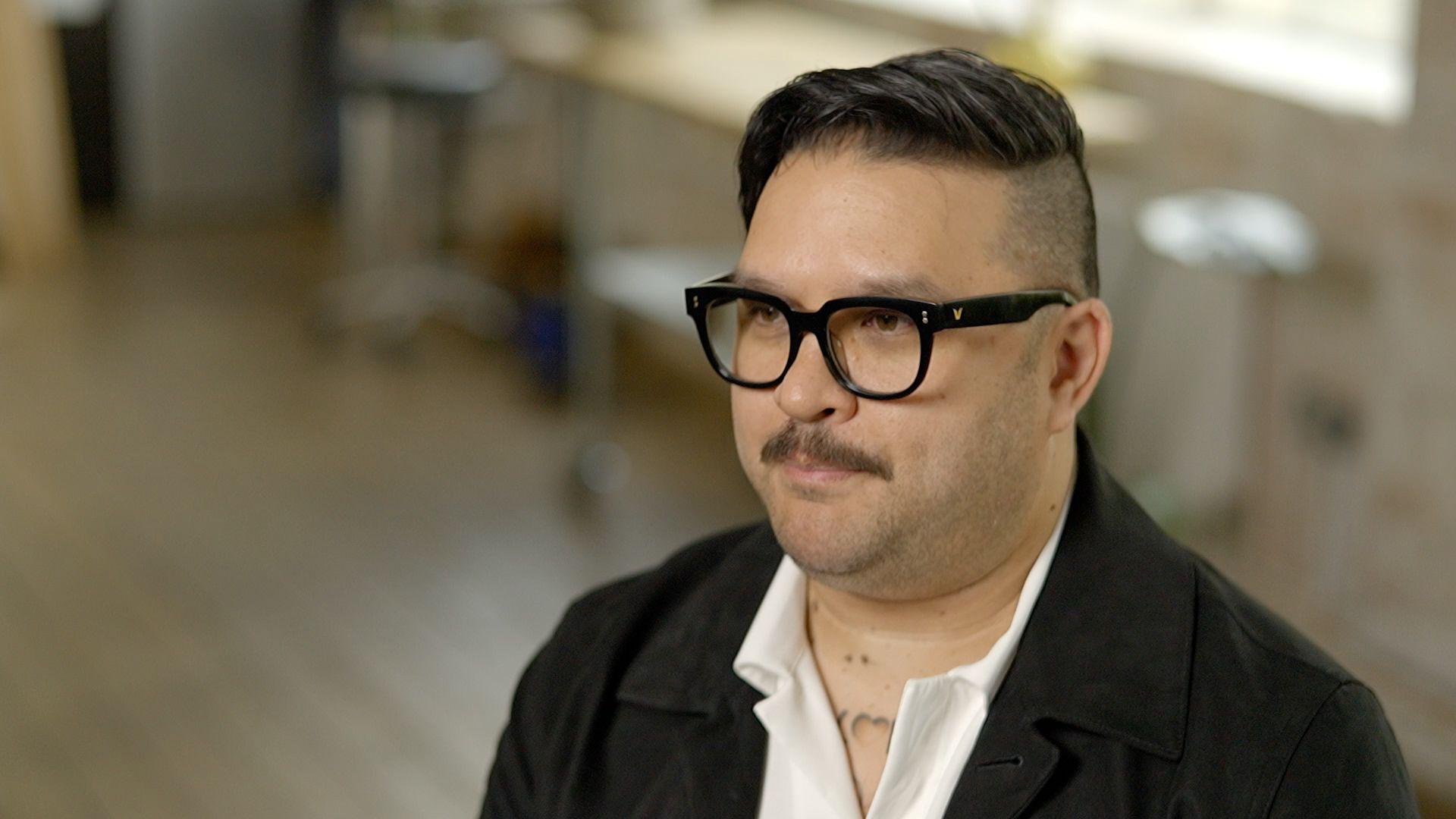 Will Hayward, a man with black-framed glasses, a moustache and dark brown hair which is shaved close to the skin on his left side and swept over the other side. He wears a dark jacket and a white shirt open at the neck.