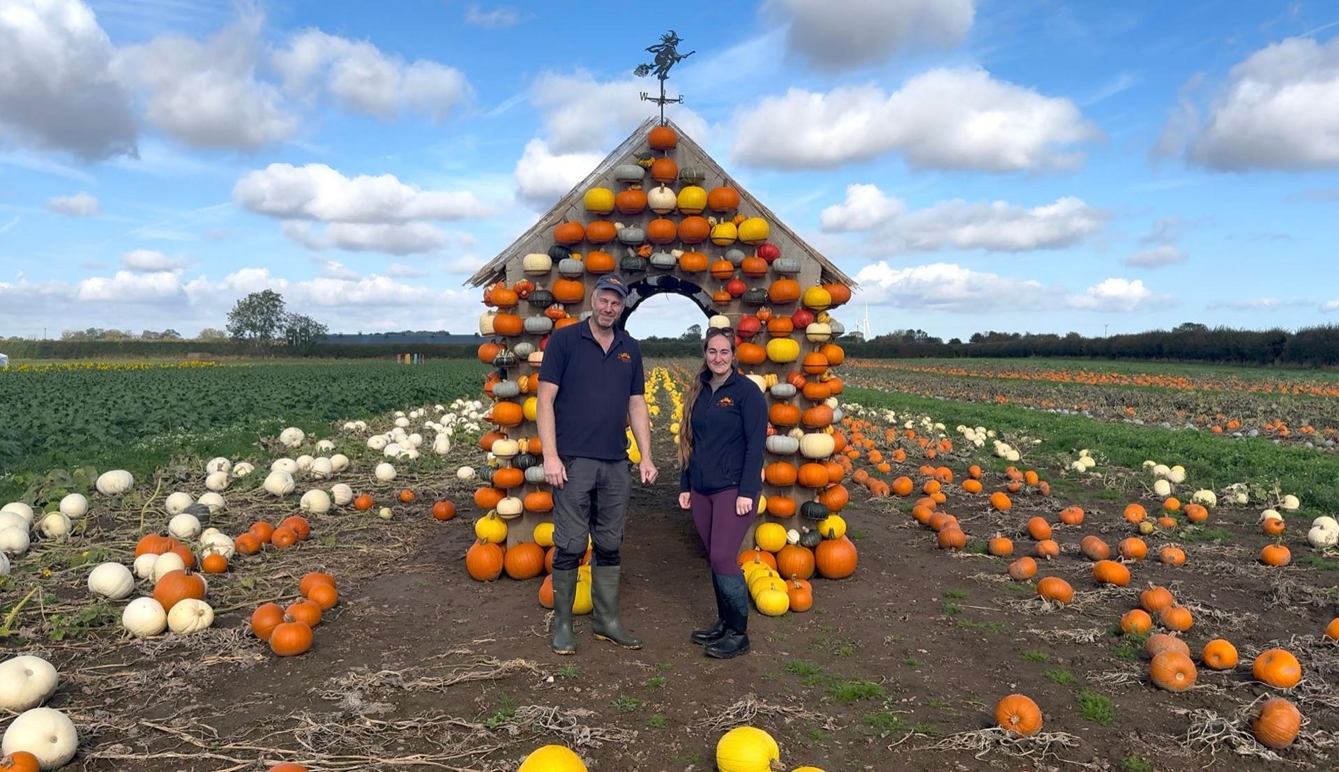 Man in blue t-shirt and blue cap and dark trousers. He is wearing wellington boots and smiling. He is stood with a woman with sunglasses on her head, blue fleece, grey top and maroon trousers. She is wearing wellington boots and smiling. There are lots of pumpkins that are white, yellow and orange in the background. There is a house shaped structure with pumpkins on it and a weather vane on top.