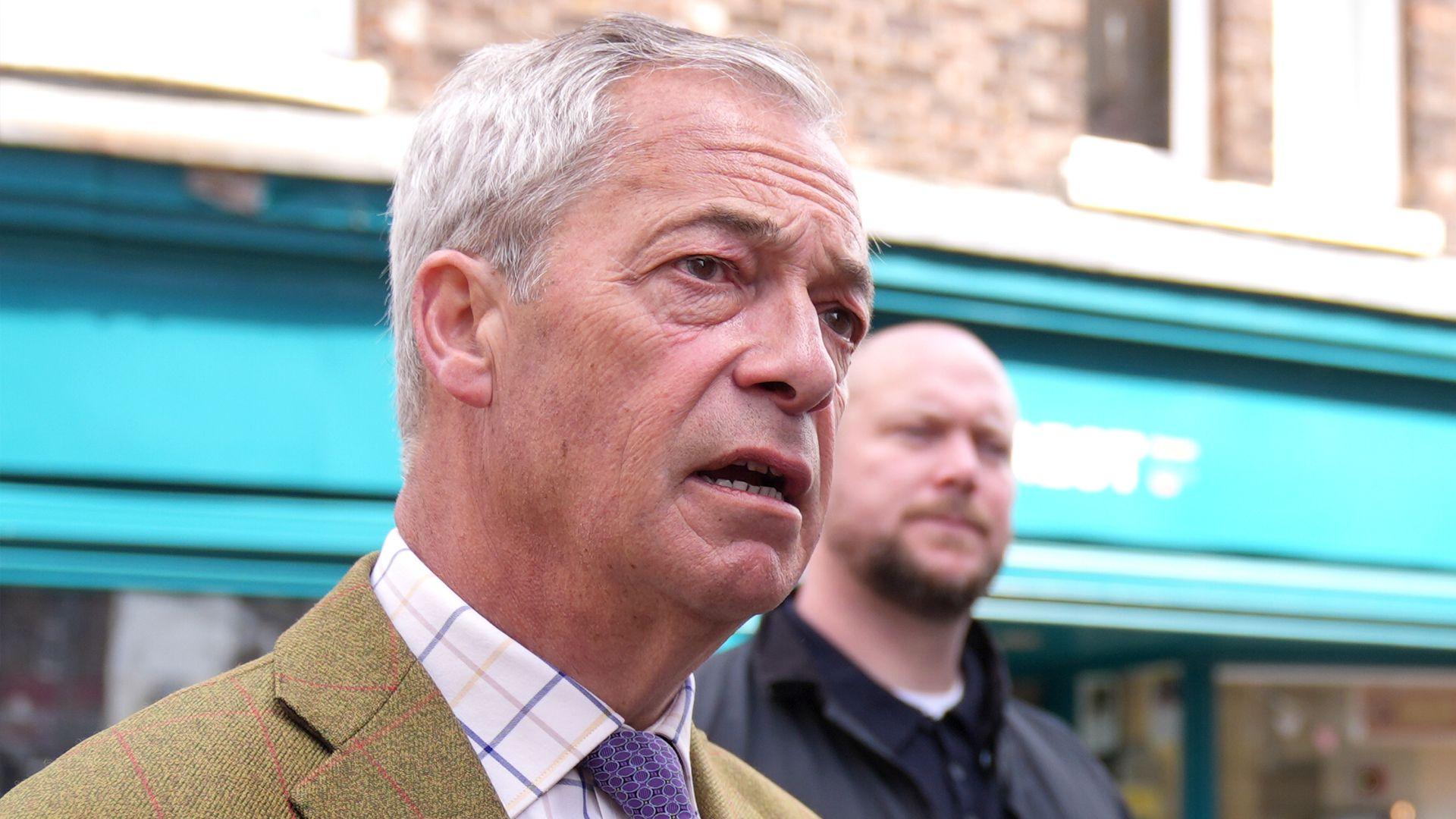 Reform UK leader Nigel Farage during a walkabout in King's Lynn, Norfolk, whilst on the campaign trail for the upcoming local elections. He is wearing a tweed jacked, checked shirt and tie and behind him stands a bald man with a beard.