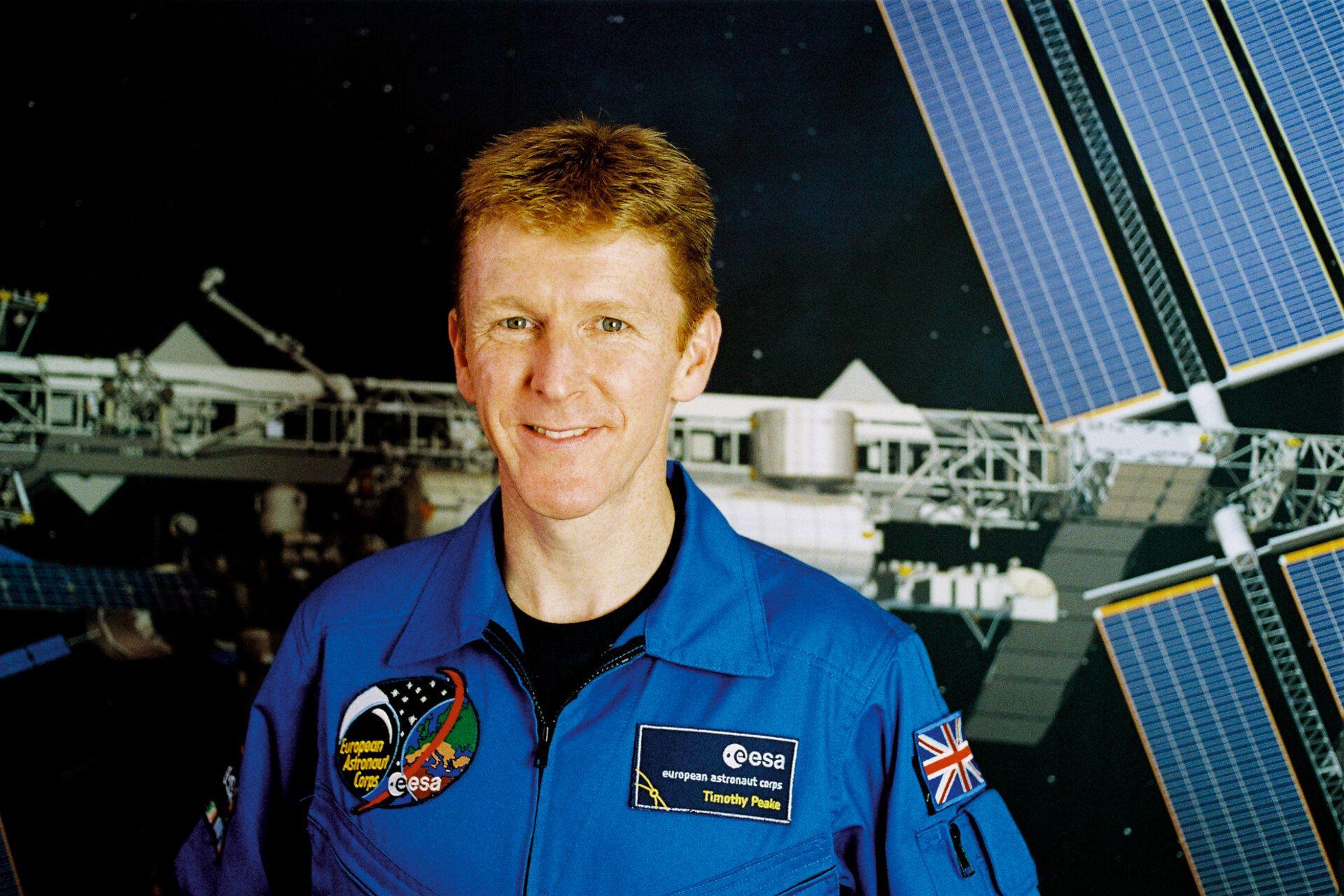 Tim Peake in a blue jump suit with two European Space Agency badges on his chest and a union flag on his left shoulder. He is standing in front of a picture of the International Space Station