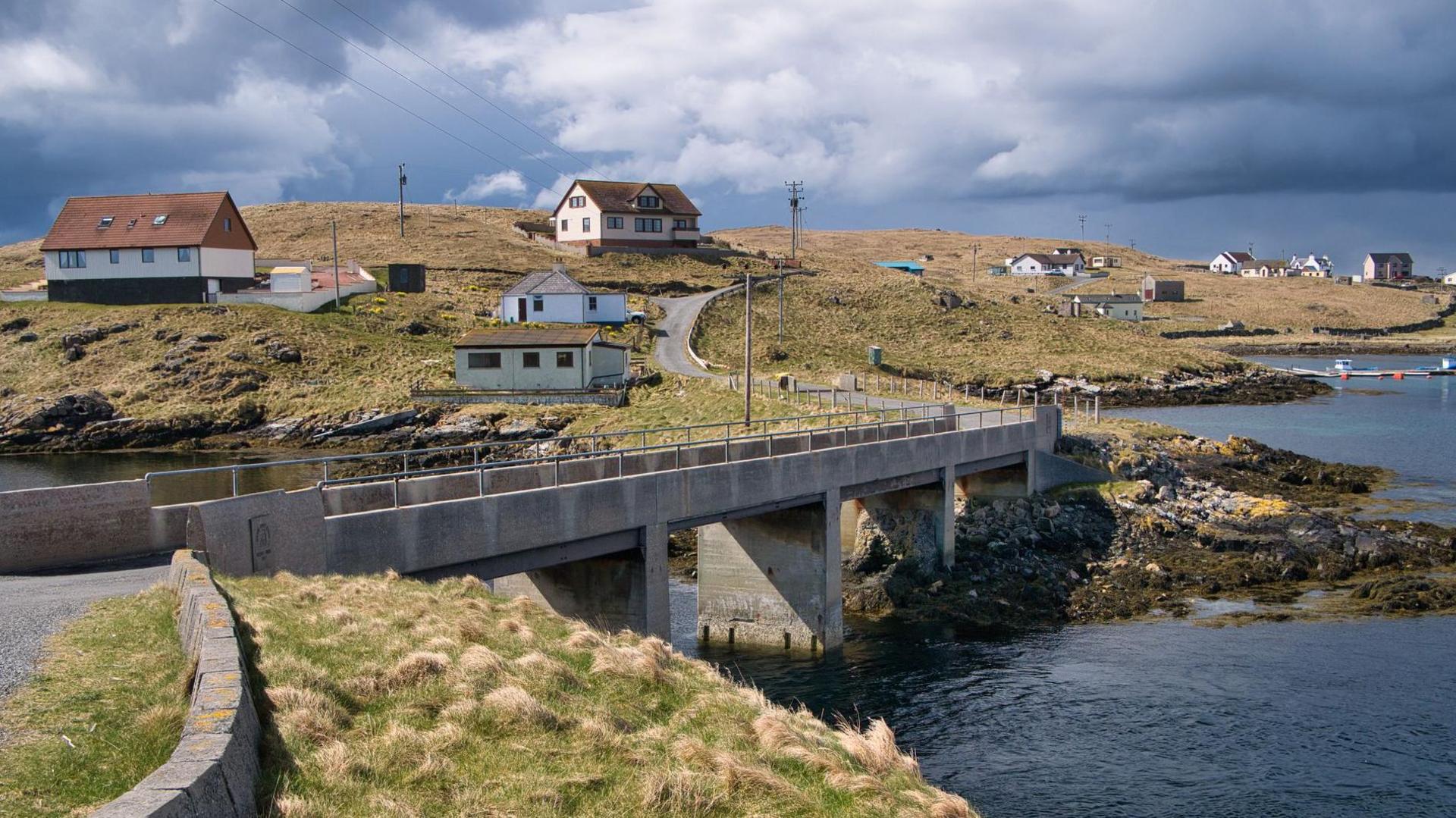 The island communities of Housay and Bruray in the island group of Out Skerries. Houses are scattered across a grassed area with a bridge at the centre of the photograph connecting the two islands and the sea on the right.