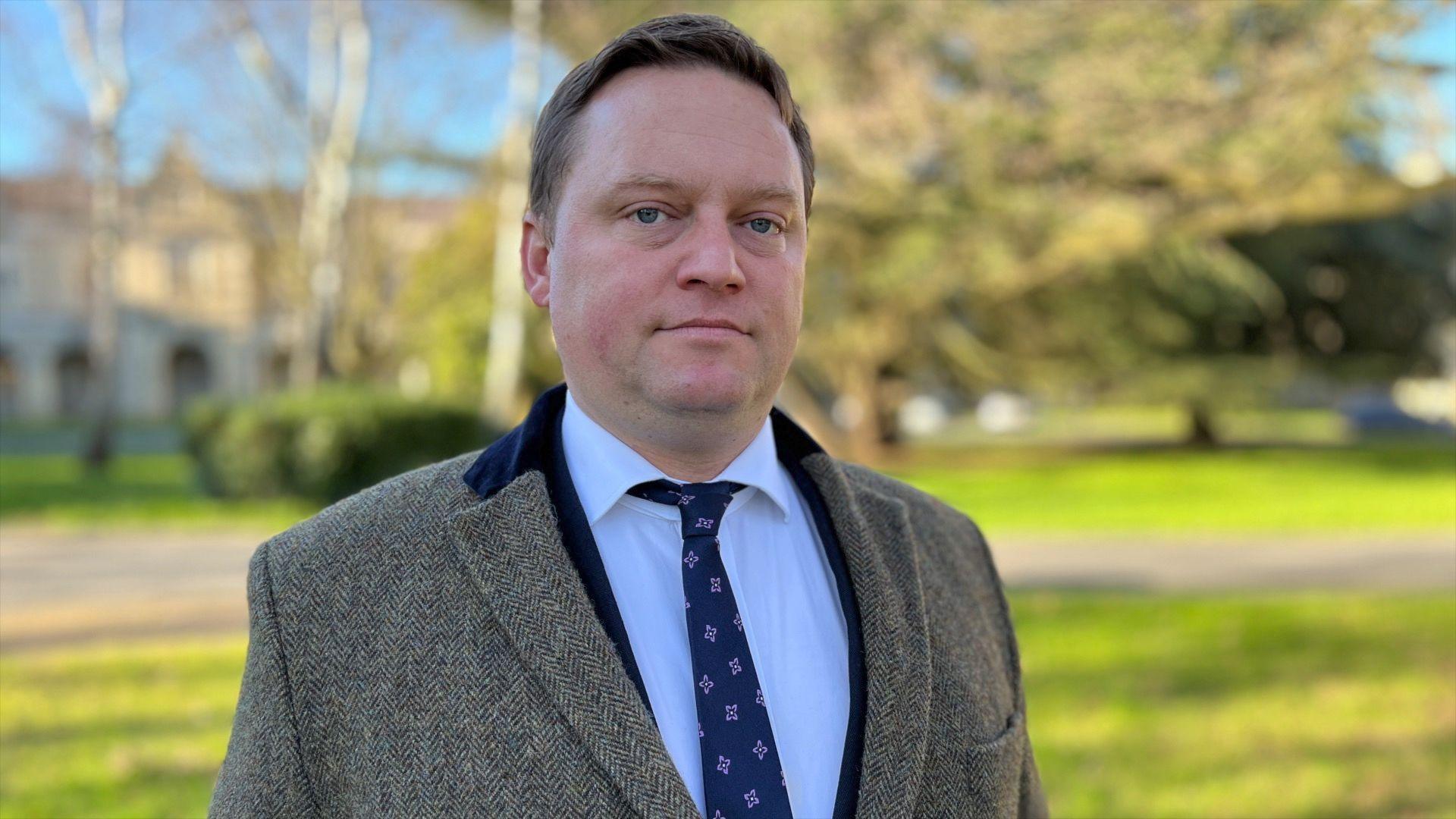 Barrister Michael Polak from Justice Abroad. He is stood in front of a blurred-out tree and building. He is wearing a white shirt, navy blue tie and suit jacket, and a tweed overcoat.