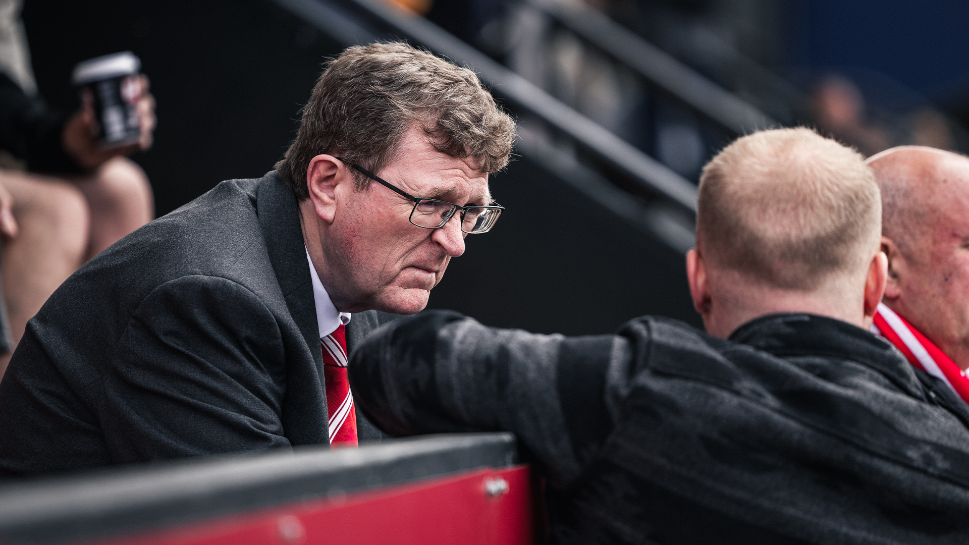 Geraint Parry in a suit leans over the barrier from his seat to talk to an unknown figure with his back to camera