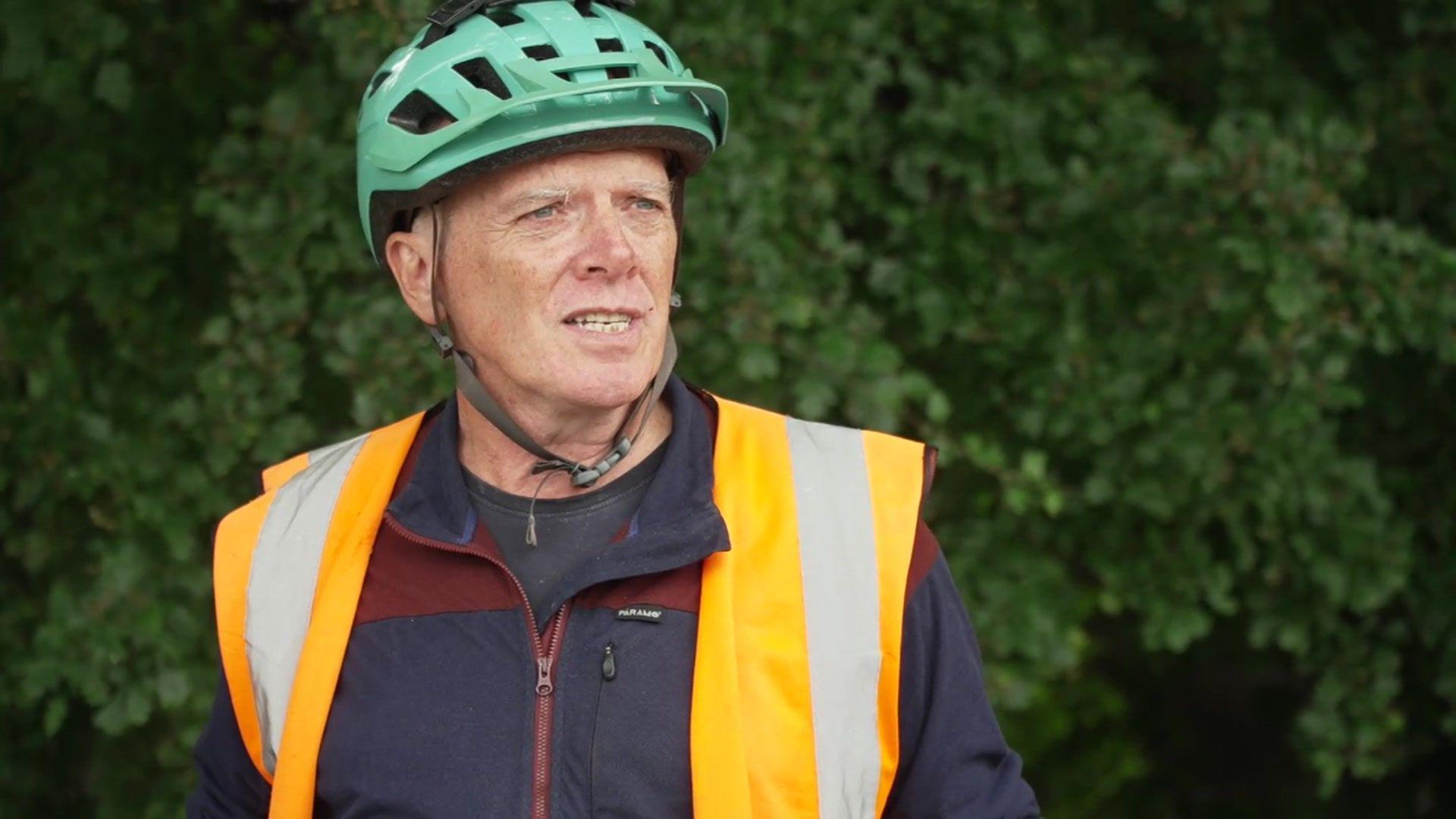 Andy Smith wearing green cycle helmet and high vis vest over a zip up jacket looking to the right of the frame, standing in front of a greenery in the background, which is out of focus.