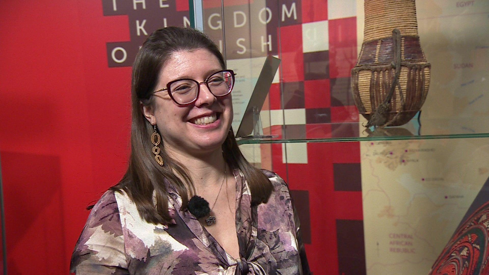 A woman with dark hair and wearing glasses and a purple blouse standing in front of a glass museum exhibit.