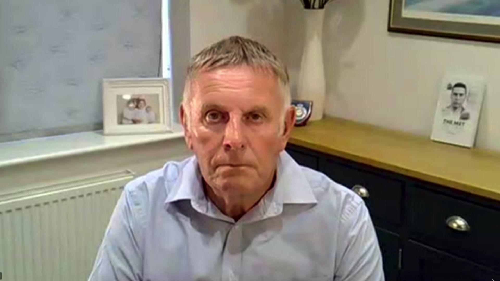 Steve Reynolds is sat in a room inside his house. He has a copy of the book over his left shoulder and a framed family photo behind his right. He has a police badge on display behind him.