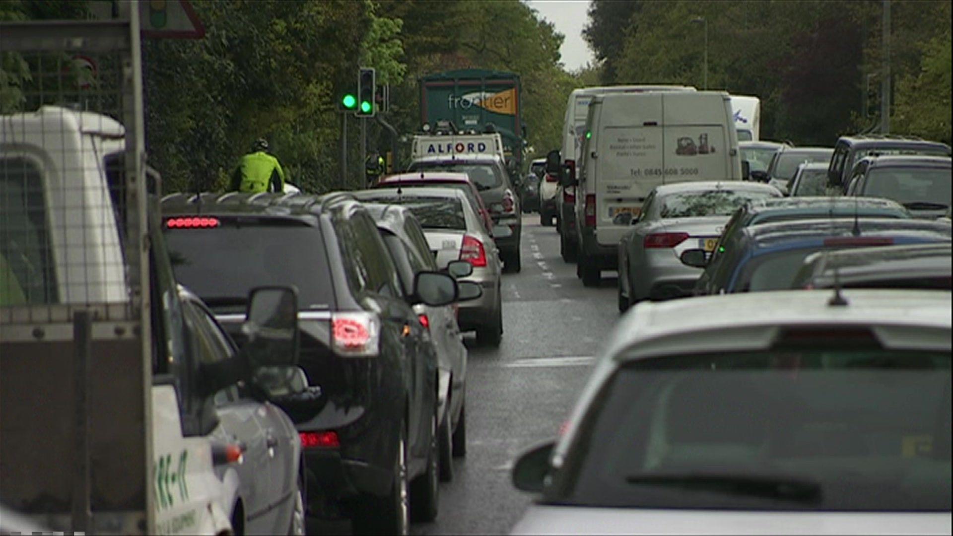 Three lanes of back-to-back traffic heading away from the camera, with green trees wither side of the road.