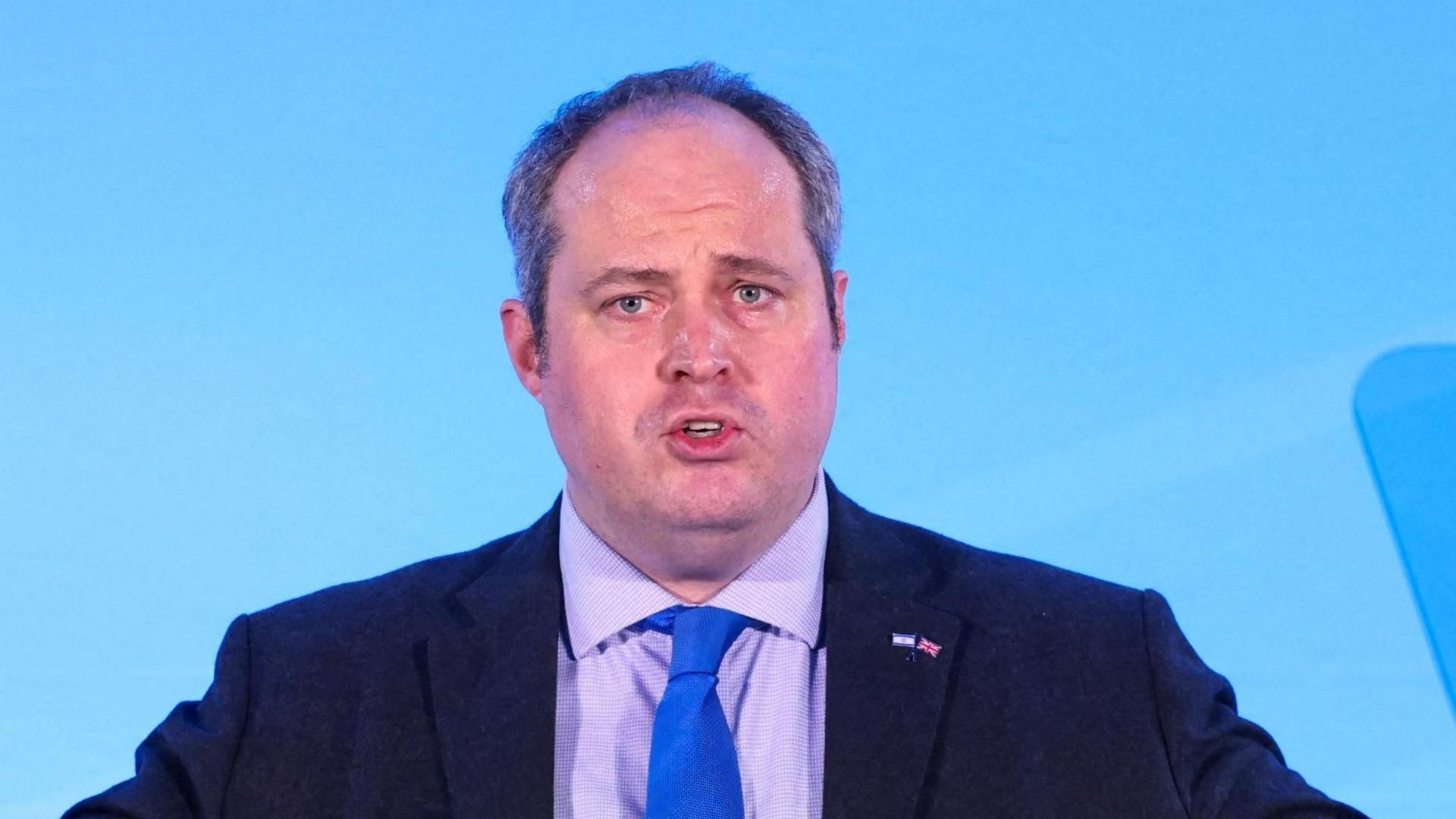 Alex Wilson is seen from the chest up, wearing a dark suit with purple shirt and blue tie. He is in the middle of giving a speech against a blue background