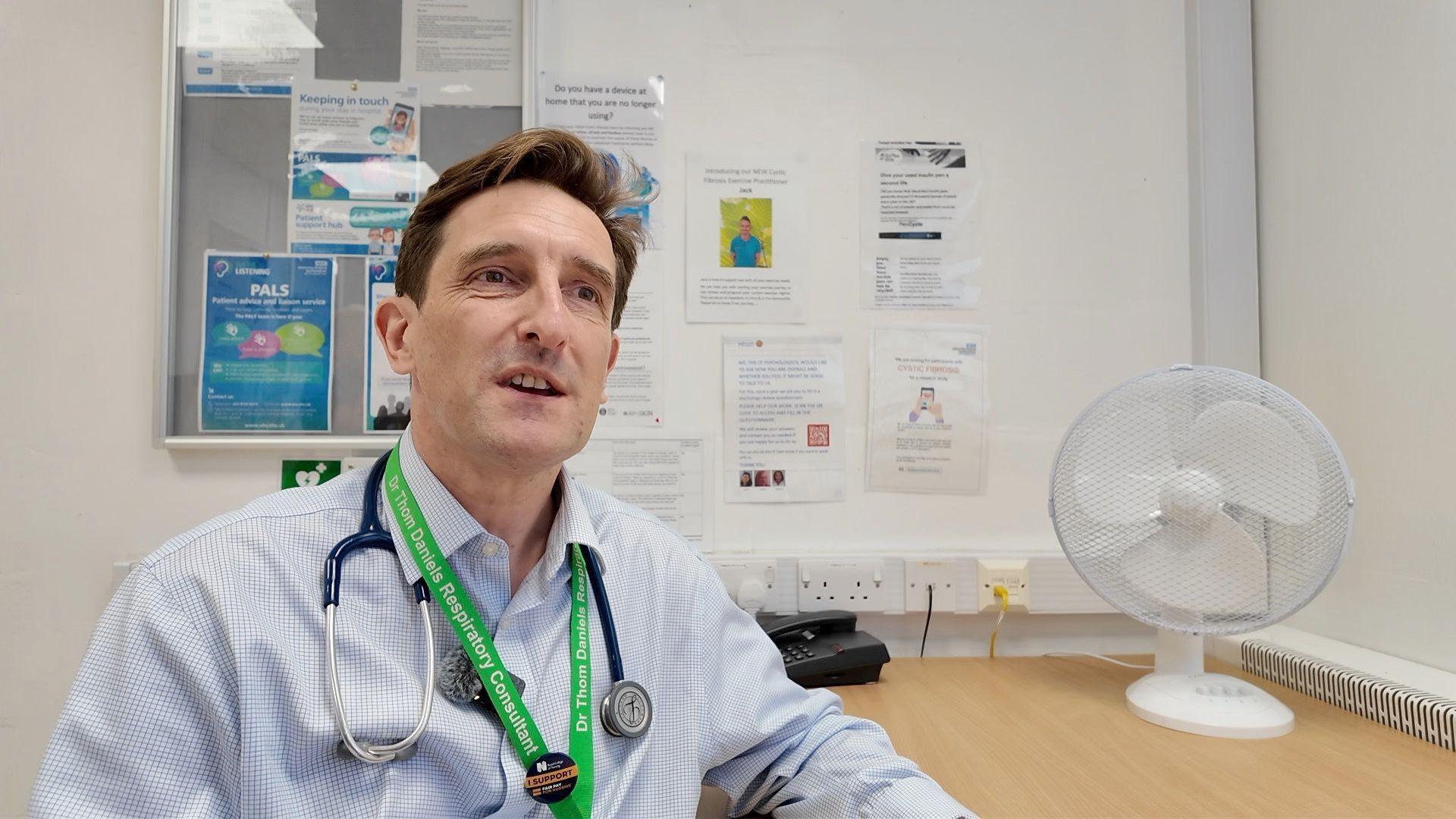 A doctor wearing a light blue shirt and with a stethoscope around his neck sits at a desk.