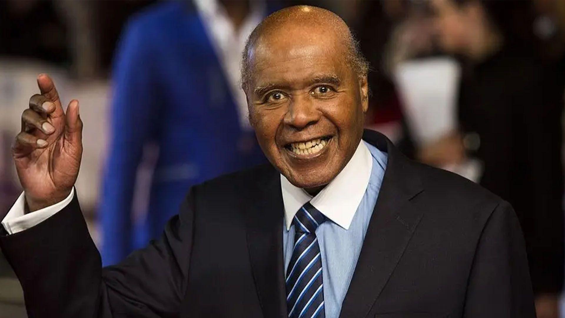 Paul Stephenson in a blue shirt, navy and pale blue striped tie and a black jacket, smiling at the camera and holding done hand up in a wave. 