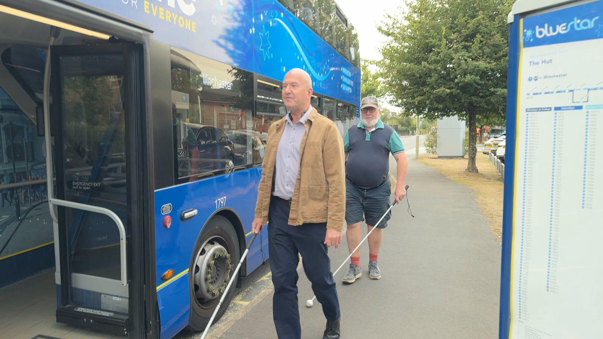Two visually impaired men walking to catch the bus. The bus is blue and the men are carrying assistance poles. 