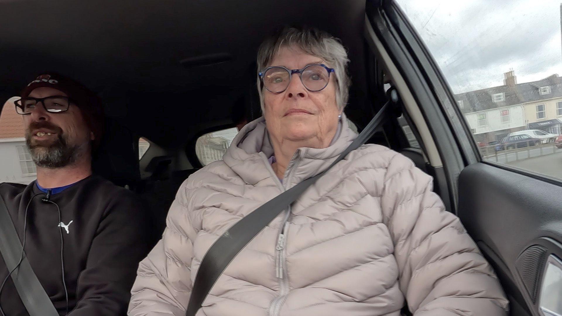 Sue Mollet strapped in a car, wearing a ribbed pink puffer jacket.
She has grey hair and blue rimmed glasses.
On her left is Chris Mollett, who is driving the car, wearing a beanie hat and a sweatshirt.