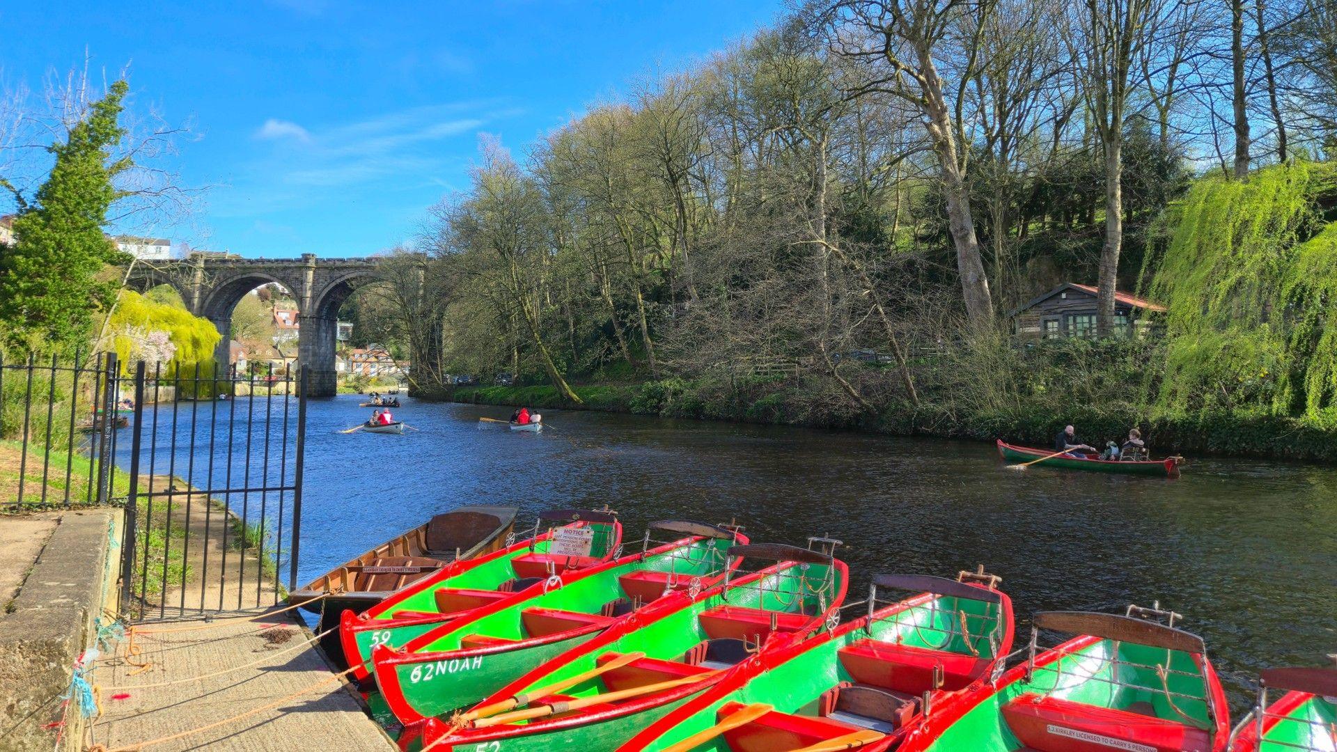 Green and red coloured rowing boats roped to the side of a tree-lined river with people boating, a railway bridge in the distance and blue sky above