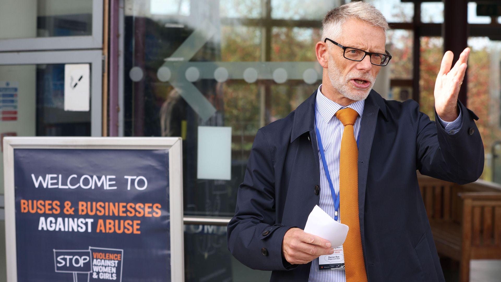 Darren Roe is standing at a bus station next to a sign saying: "Welcome to Buses and Businesses Against Abuse." He has greying, short hair and a beard and moustache. He is wearing dark-rimmed spectacles, a blue jacket, blue striped shirt and an orange tie. His left hand is raised as he addresses onlookers.