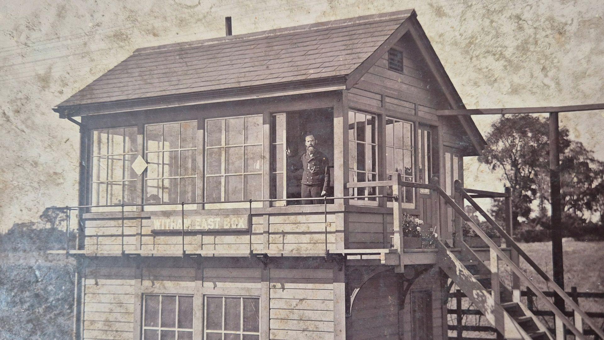 A black and white image shows the Witham East railway signal box. It is constructed from wood panelling and has several large windows. At the entrance stands Ben Sainty in his signalman uniform.