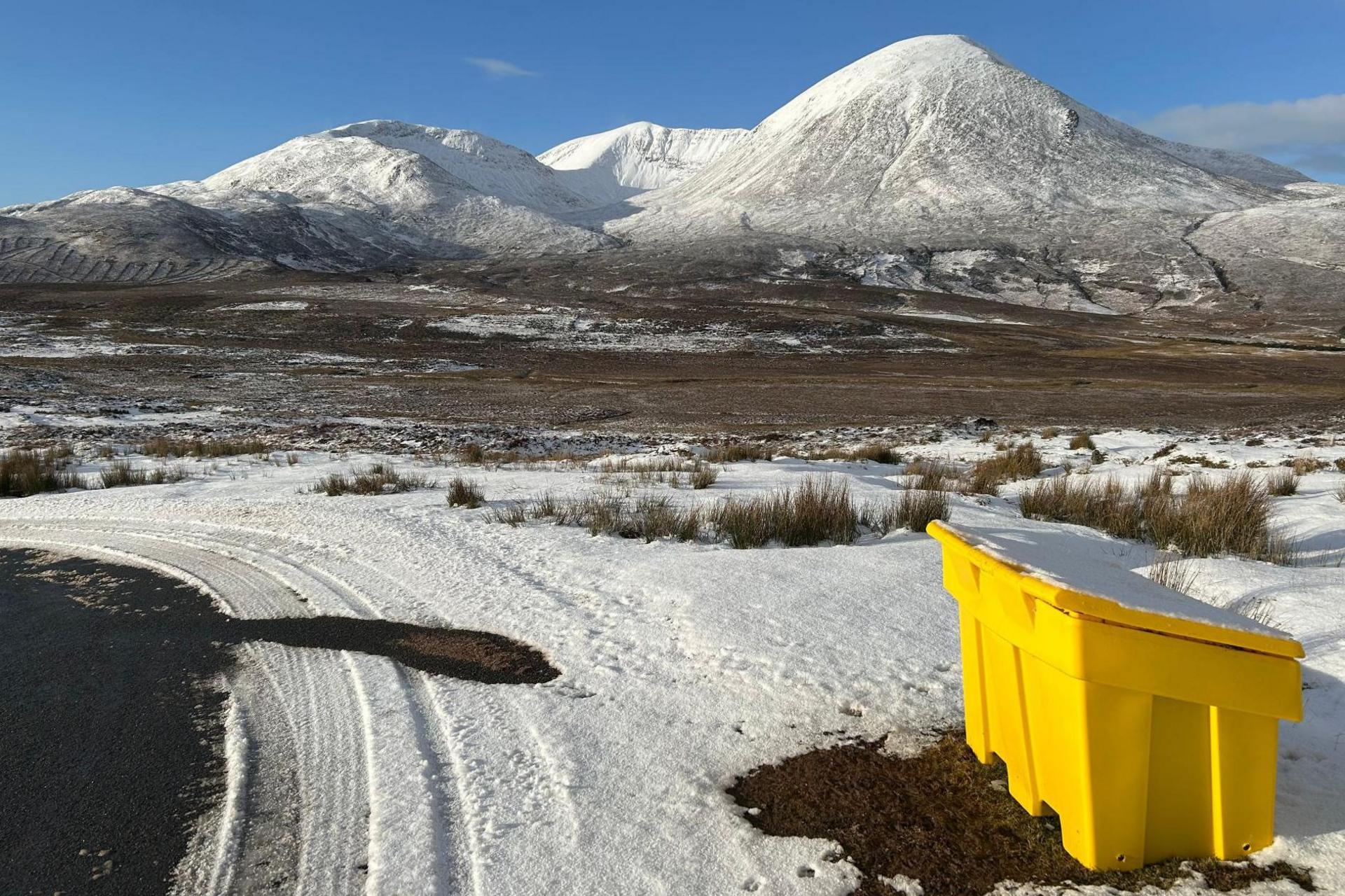 There is a bright yellow road salt bin on a snowy road verge. In the distance are mountains of Skye under a blue sky.