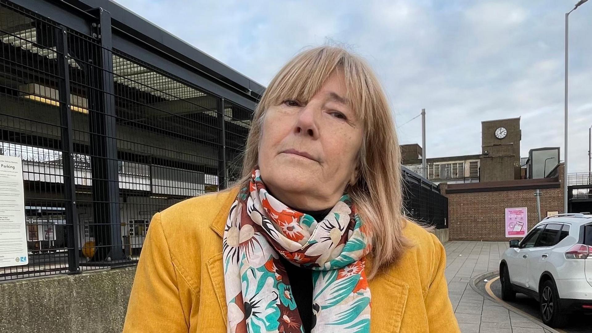 Looking direct to camera, Marie McCormick is standing outside Luton Station. She is wearing a yellow corduroy jacket and a flower print silk scarf. She has shoulder length blond hair. Her head is slightly tilted and twisted due to her living with dystonia. 