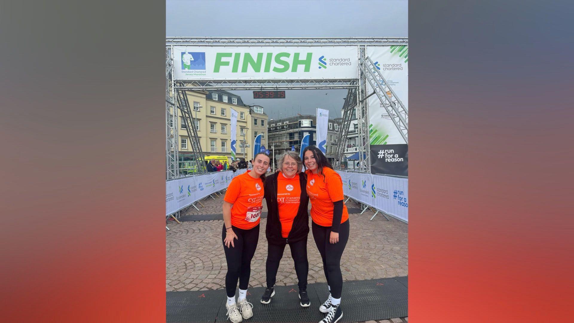 All three smile at the camera at the finish line of the Jersey Marathon. They're wearing bright orange tops and black running leggings.