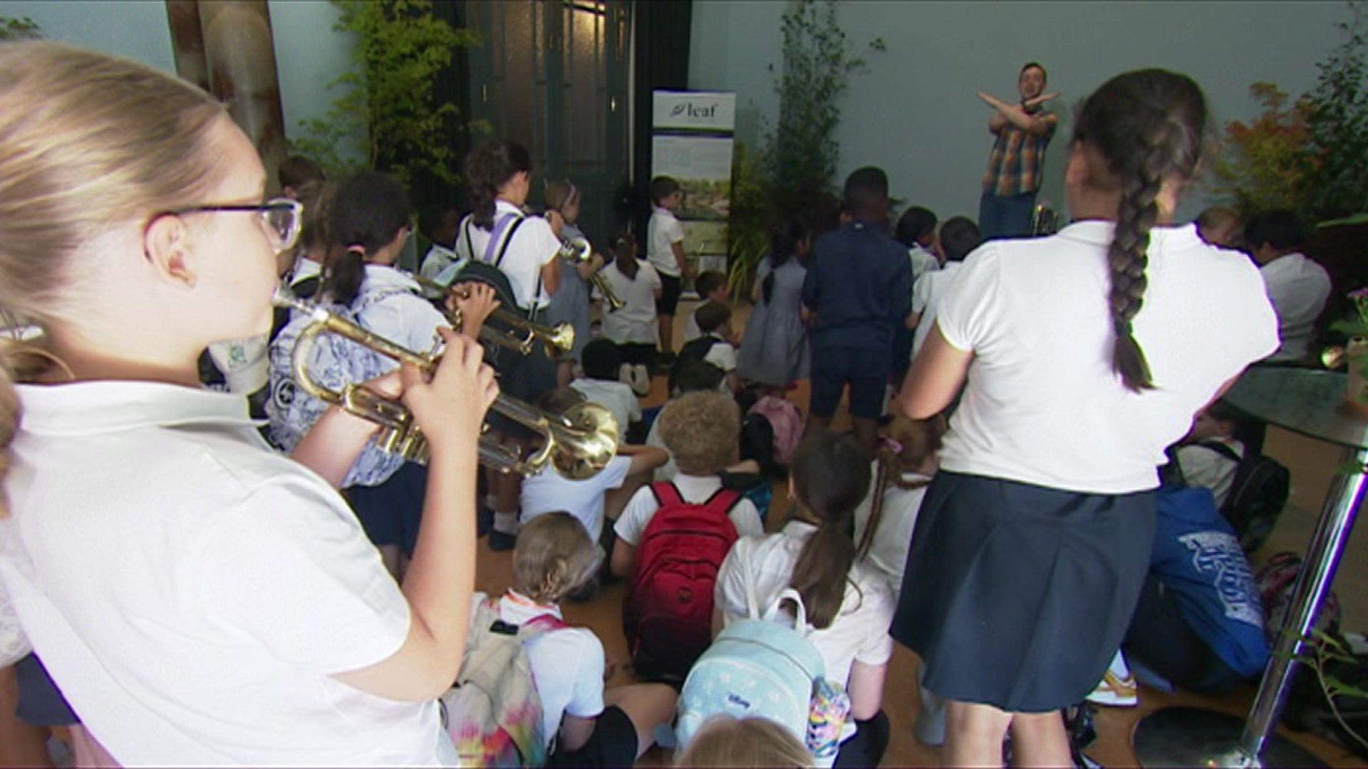 A group of schoolchildren in uniform play trumpets as a man conducts them at the front of the room