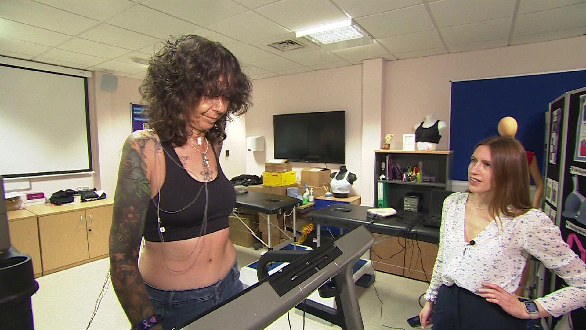 Celeste, a lady with brown curly hair, walks on the treadmill in a bra. She has white monitors on her. Whilst the clinical research fellow of the study, Amy in a white blouse, looks on. 