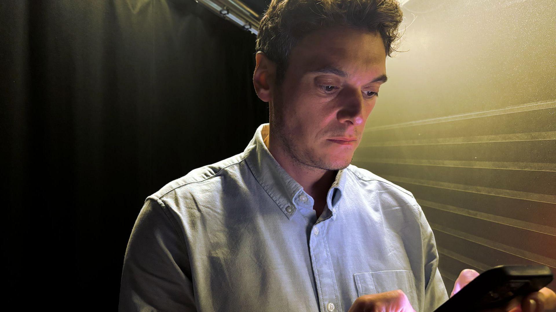 Joe Tidy looking down at a phone. He has short brown hair and is wearing a light blue shirt.
