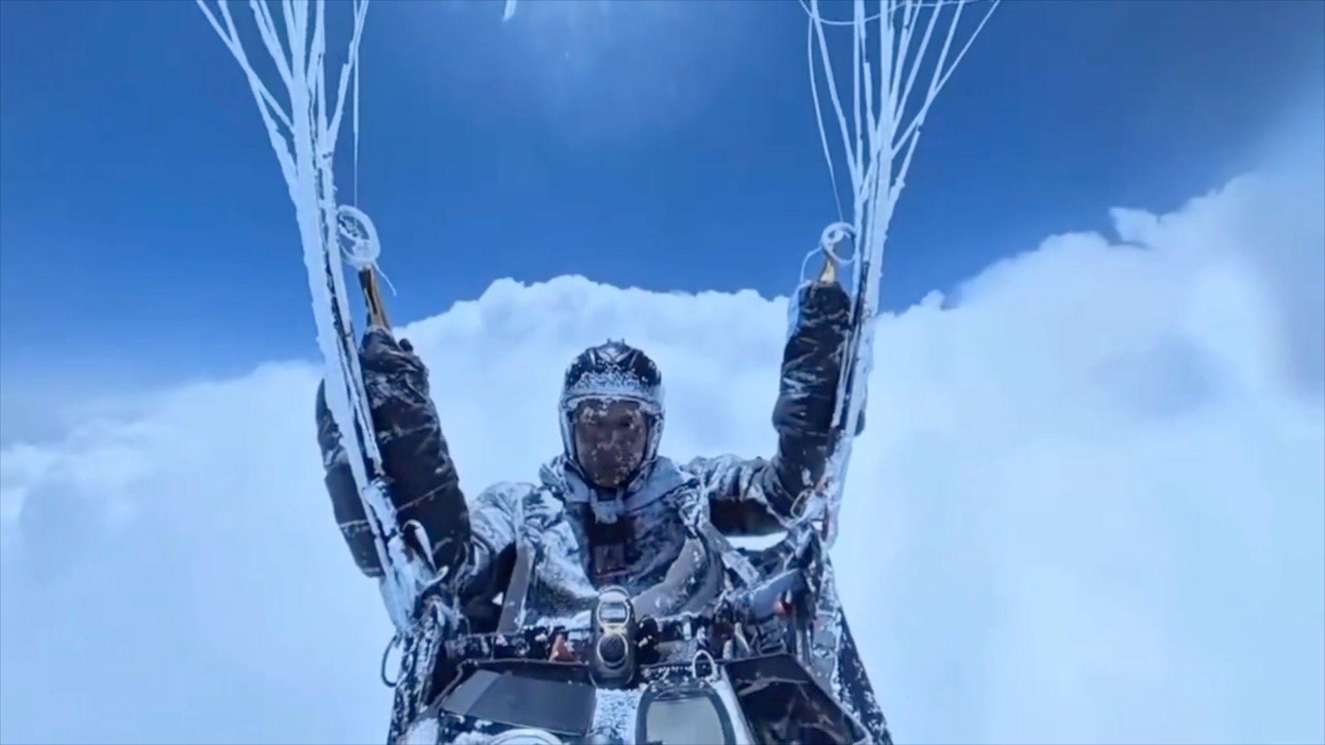 A paraglider covered in ice, wearing a helmet, goggles, big gloves and equipment, holding onto the ropes, floats in front of a blue sky and white clouds