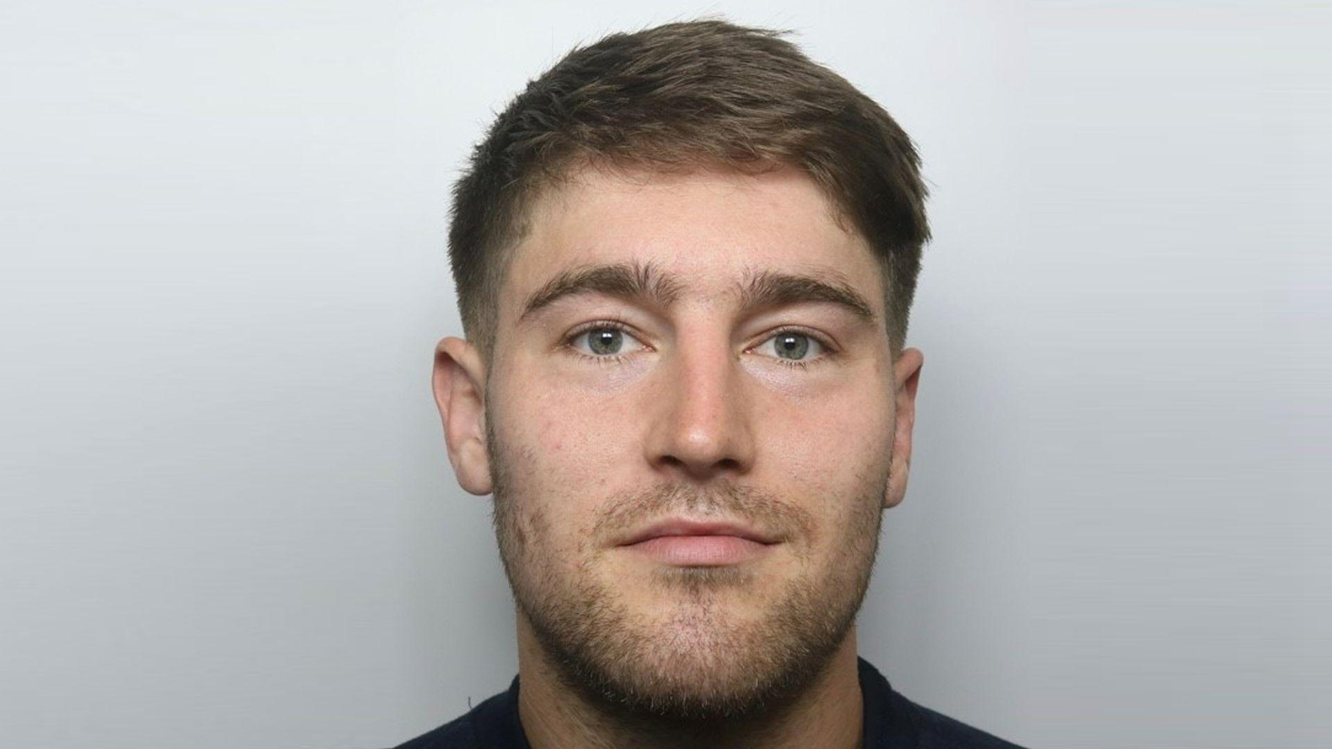 A man with short brown hair and stubble looks straight at the camera and smiles slightly in a police mugshot.