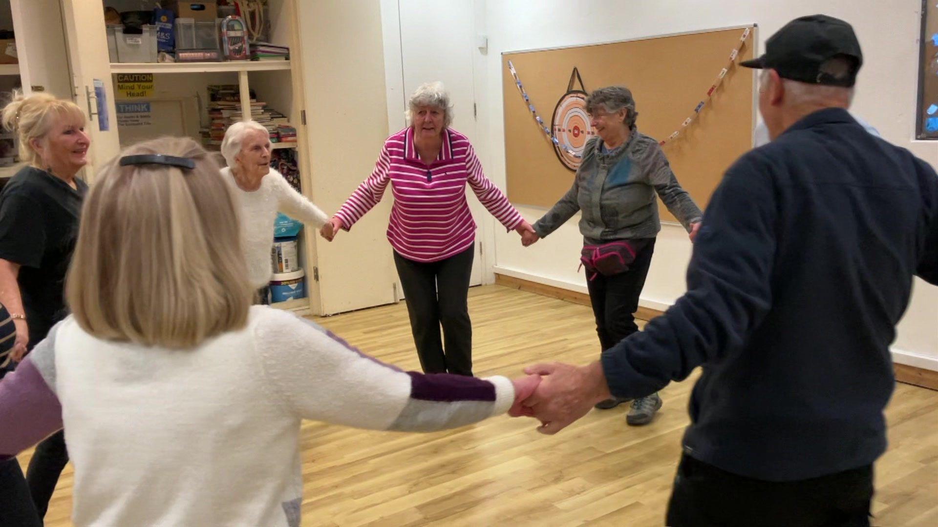 Photo of group of elderly people in a circle during a music and dancing session, holding hands and smiling.